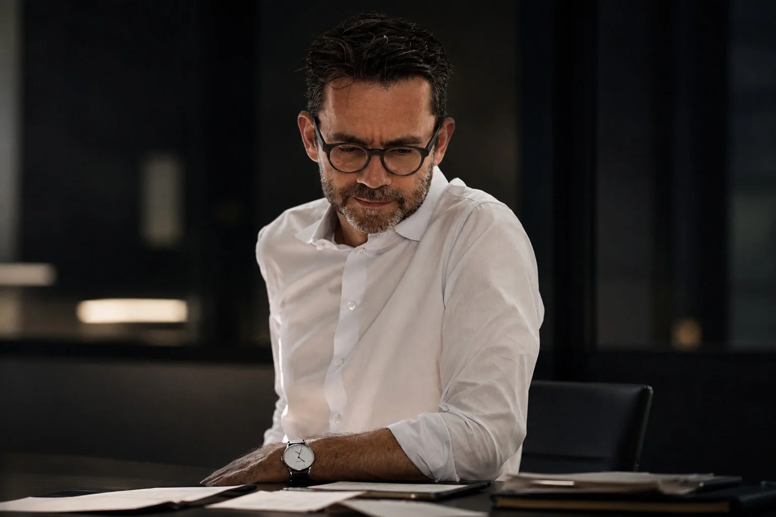 Guido Schommer mit Brille und grauem Bart sitzt an einem Schreibtisch, trägt ein weißes Hemd und schaut nach unten auf Papiere.
