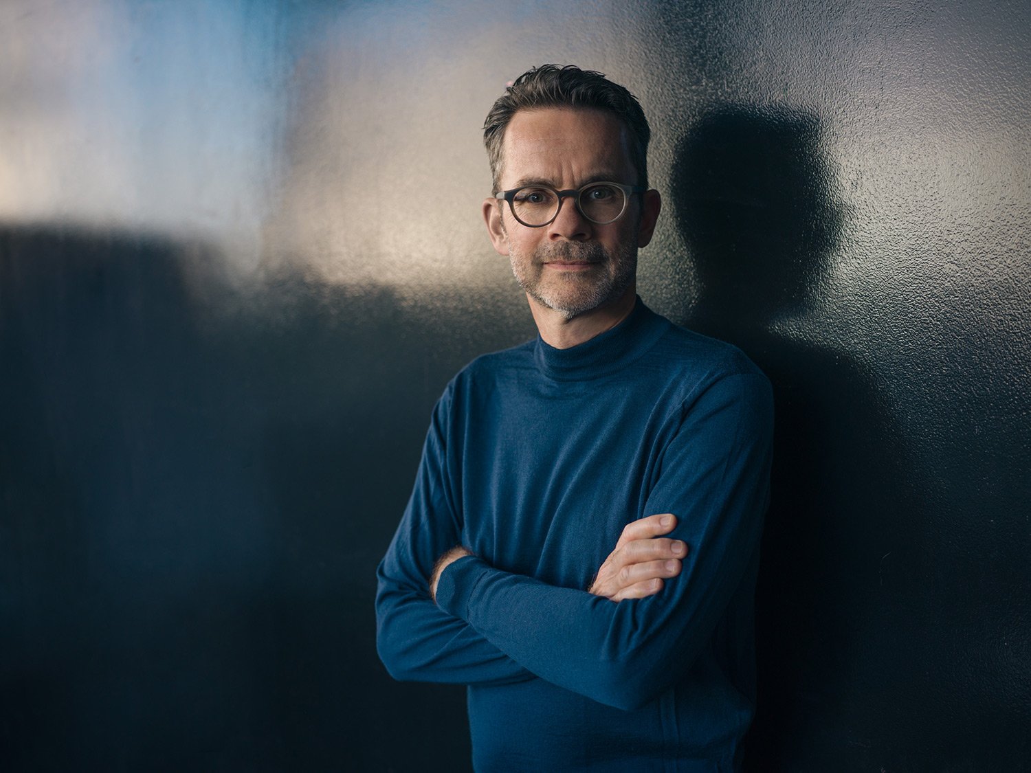 Guido Schommer in einem blauen Pullover lehnt an einer dunklen Wand und schaut nach vorne.