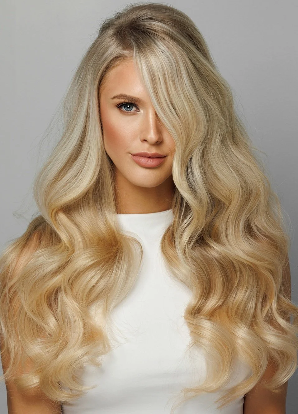 A woman with long, wavy blonde hair and blue eyes, wearing a white top, posing against a plain gray background. Full or partial Highlights available at Salon Amoura near the University of Central Florida.