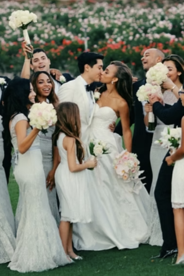 A wedding celebration with the bride and groom kissing, surrounded by friends and family holding flowers outdoors. 