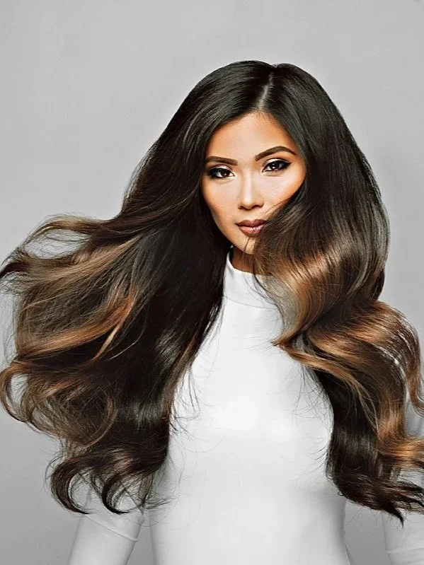 A woman with long, voluminous dark brown hair with caramel highlights, wearing a white top, looking at the camera against a plain gray background. Balayage hair done on Harana  by Salon Amoura near UCF avalon park bithlo