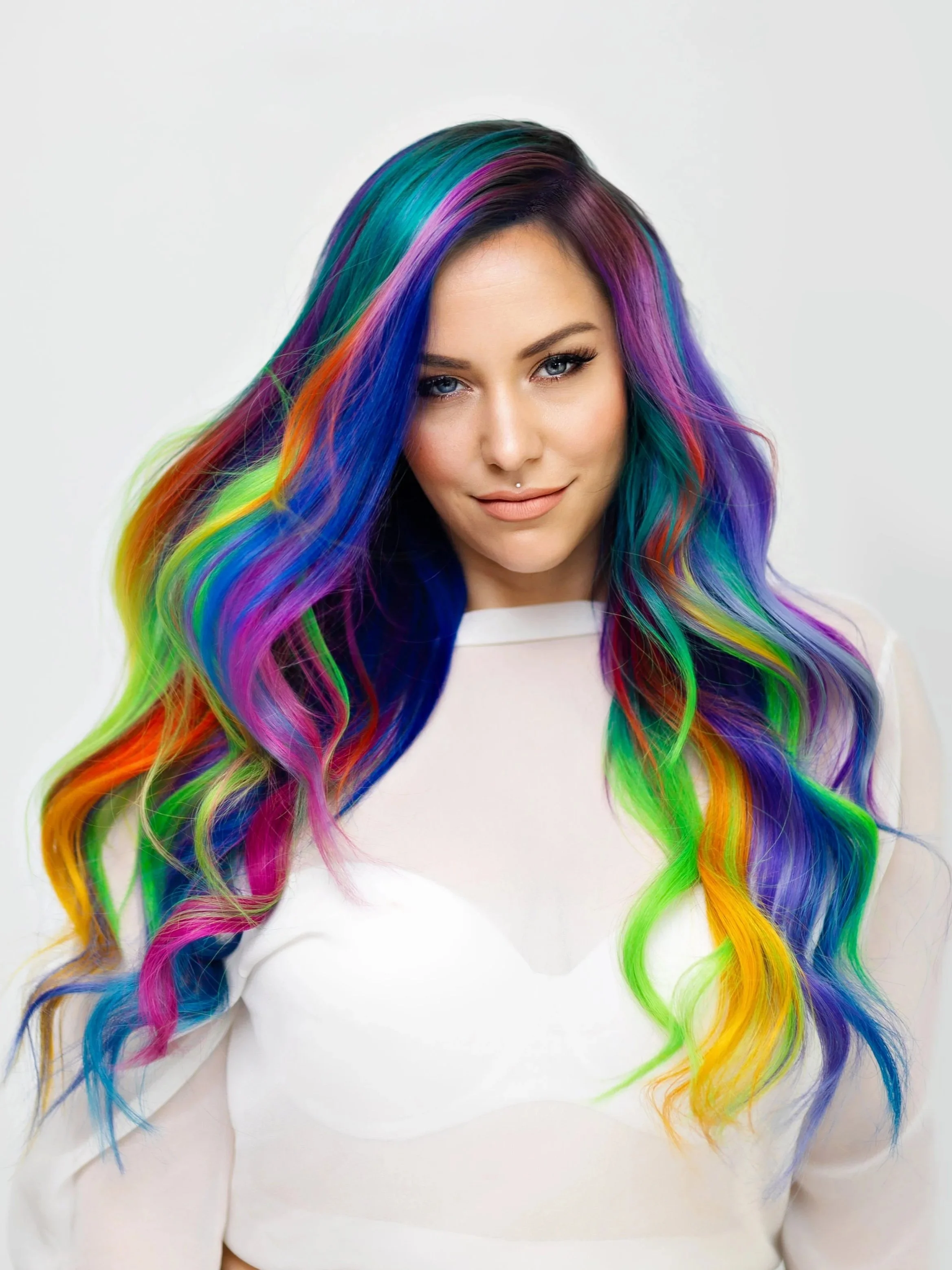 Close-up of a woman with long rainbow-colored hair, wearing a white top, and a subtle smile.