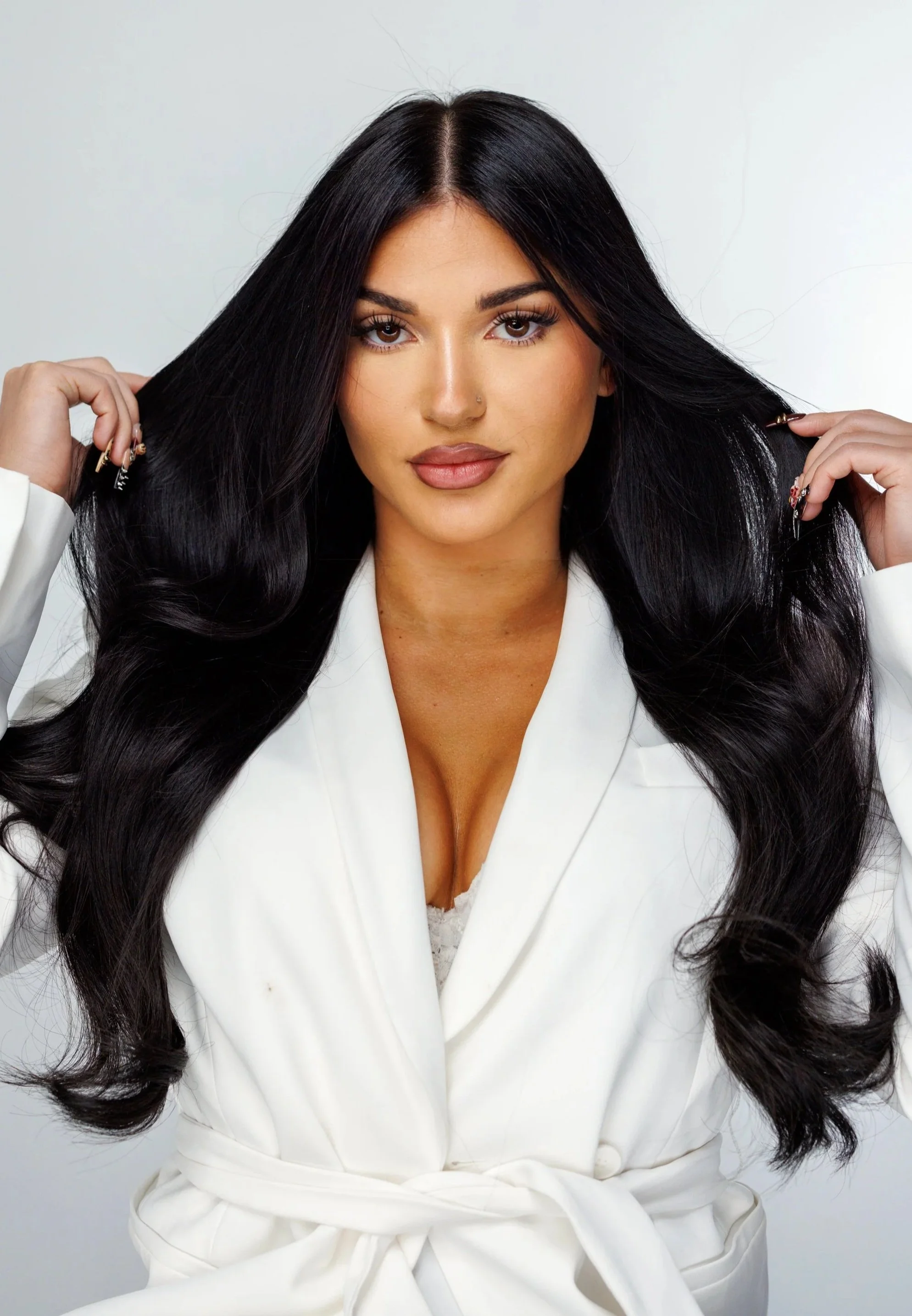 A woman with long black hair, wearing a white blazer, holding her hair with both hands, against a plain light gray background.