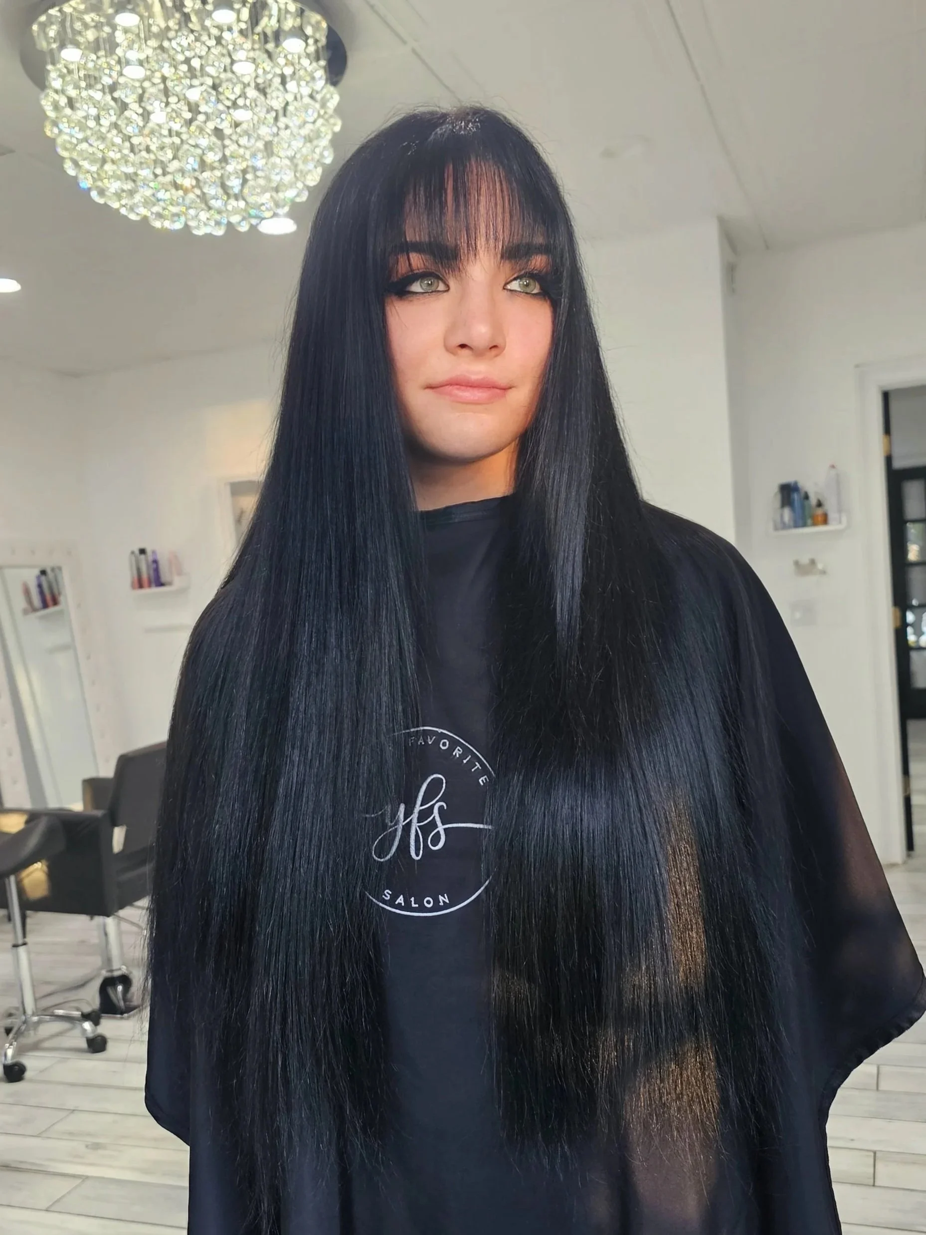 Woman with long black hair and bangs sitting in a salon, wearing a black salon cape, with salon chairs and mirrors in the background.