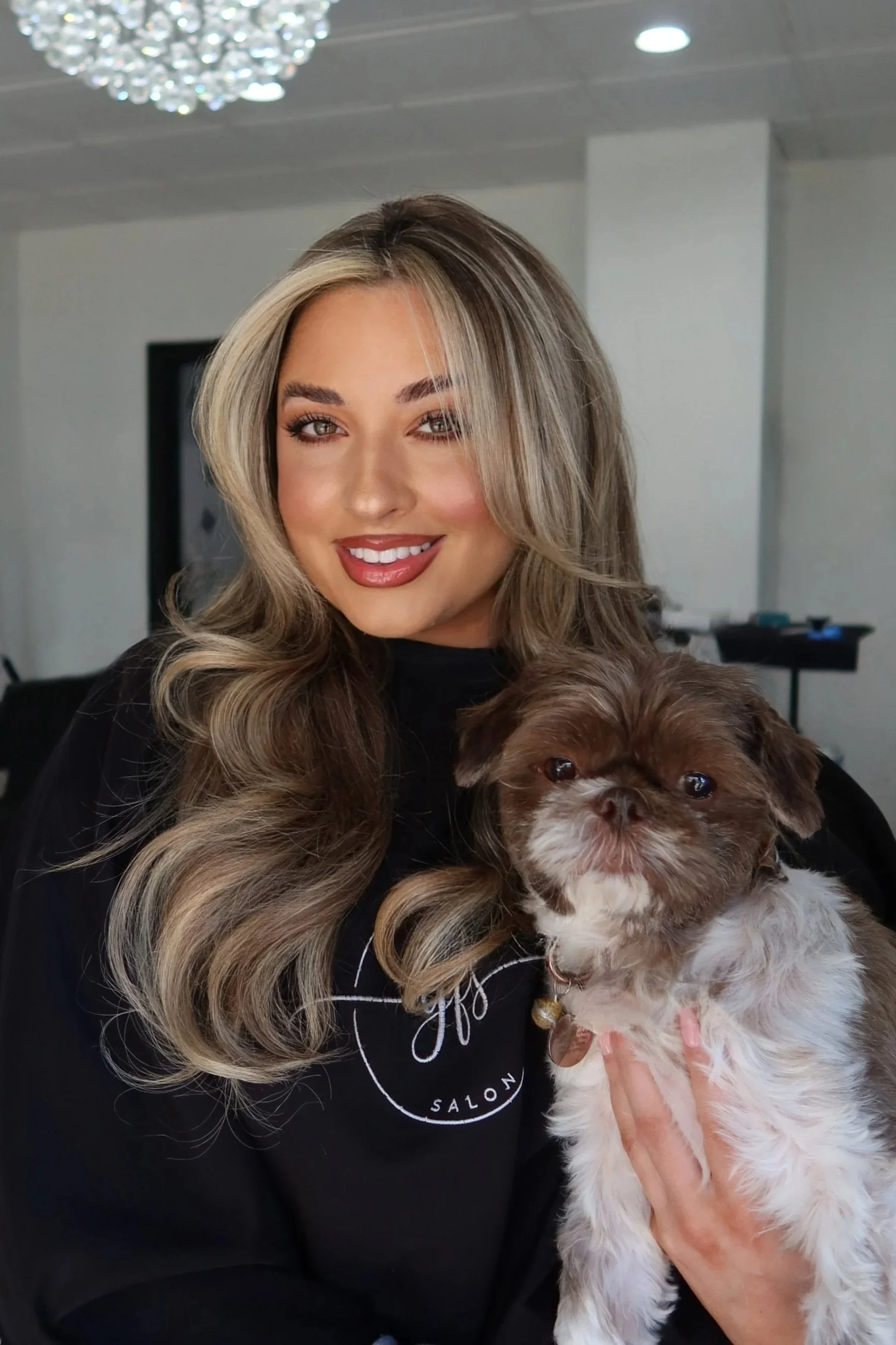 A woman with long, wavy blonde hair, wearing makeup, and smiling, holding a small brown and white dog in her arms inside a modern room with white walls, black-framed windows, and a chandelier. Bynicolemaddie gets 1 row of hair extensions and color