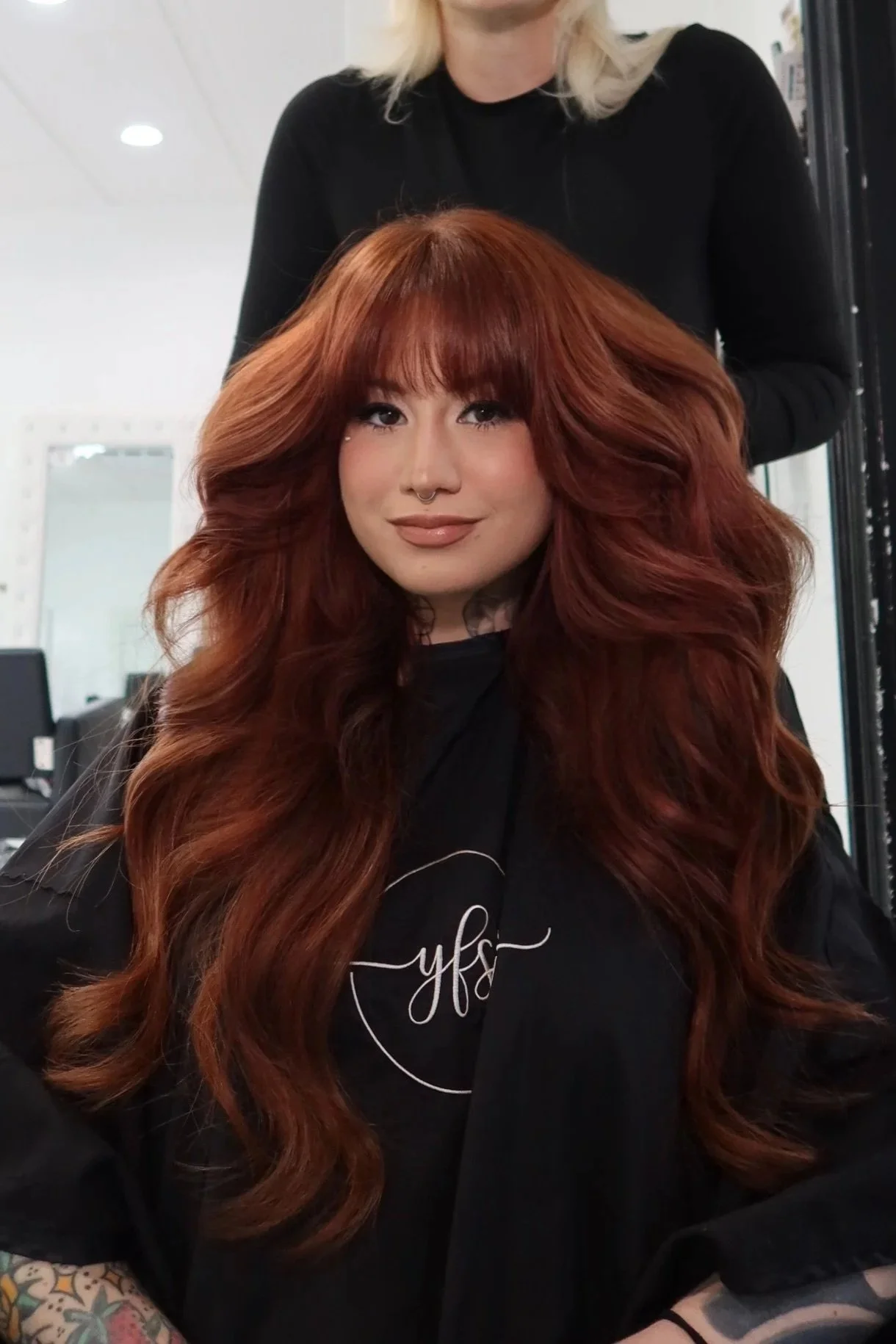 A woman with long, curly red hair, makeup, and a septum piercing, sitting in a salon chair with a stylist behind her. The woman is wearing a black top with a graphic design. Red Hair cowboy copper done at Salon Amoura Orlando
