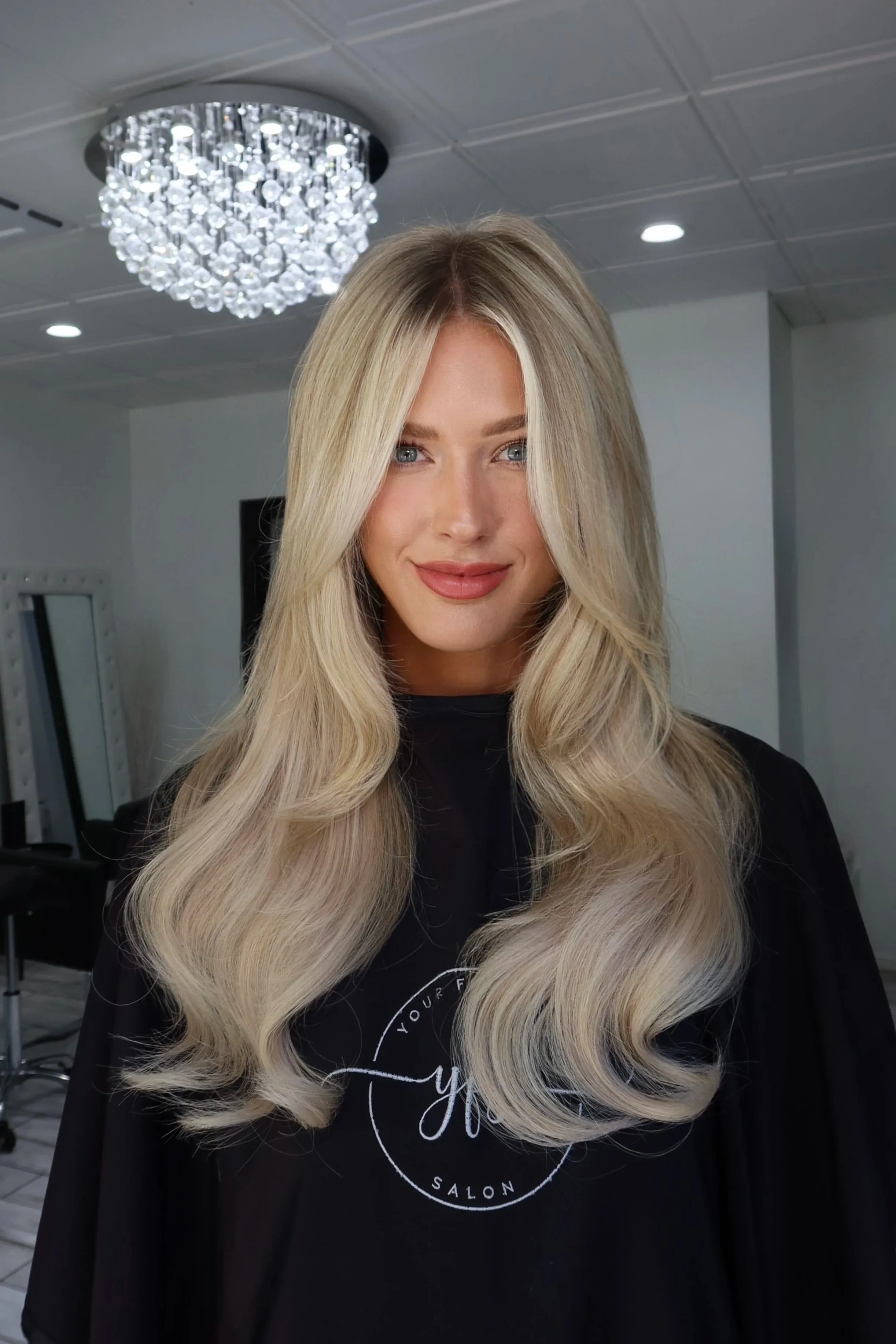 A young woman with long, wavy blonde hair and light skin, smiling at the camera, standing in a salon with a chandelier and salon equipment visible in the background. Platinum blonde done by Salon Amoura near UCF