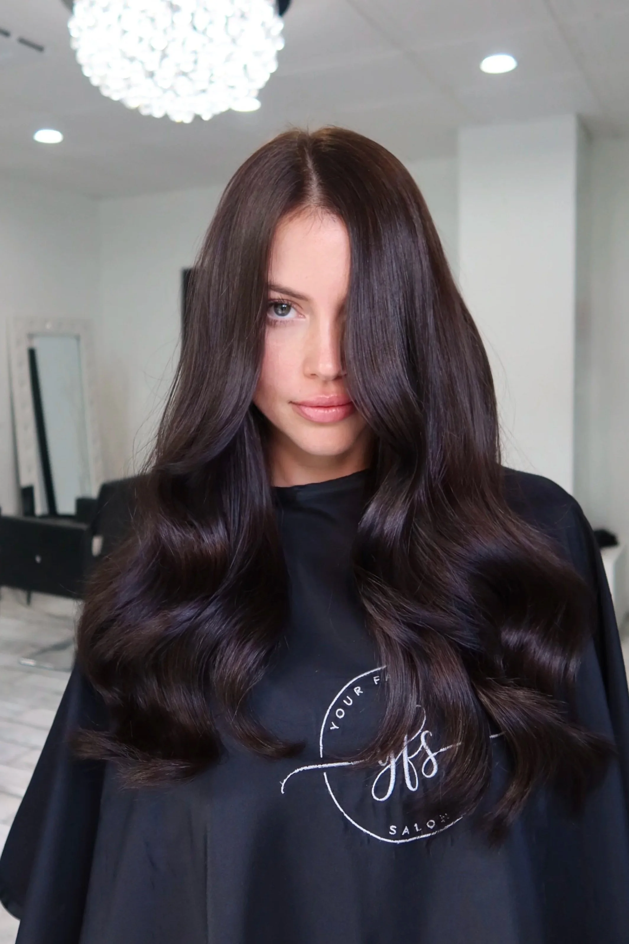 Woman with long, dark, wavy hair sitting in a salon. Bright lighting and mirrors are visible in the background. Chocolate Brown hair and 1 row of hair extensions done by Salon Amoura near UCF