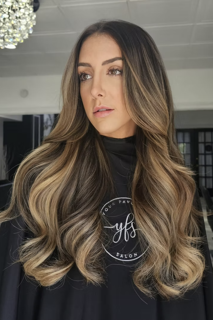 A woman with long, wavy, ombré hair in shades of brown and blonde sits in a salon. AEW Britt Baker gets 2 rows of hair extensions done by Salon Amoura near UCF orlando florida bithlo avalon park waterford lakes