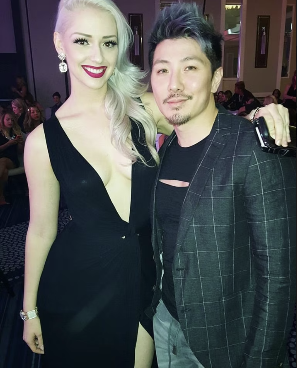 A woman with platinum blonde hair and a black dress poses next to a man in a black shirt and plaid blazer at an indoor event, with a group of people in the background.
