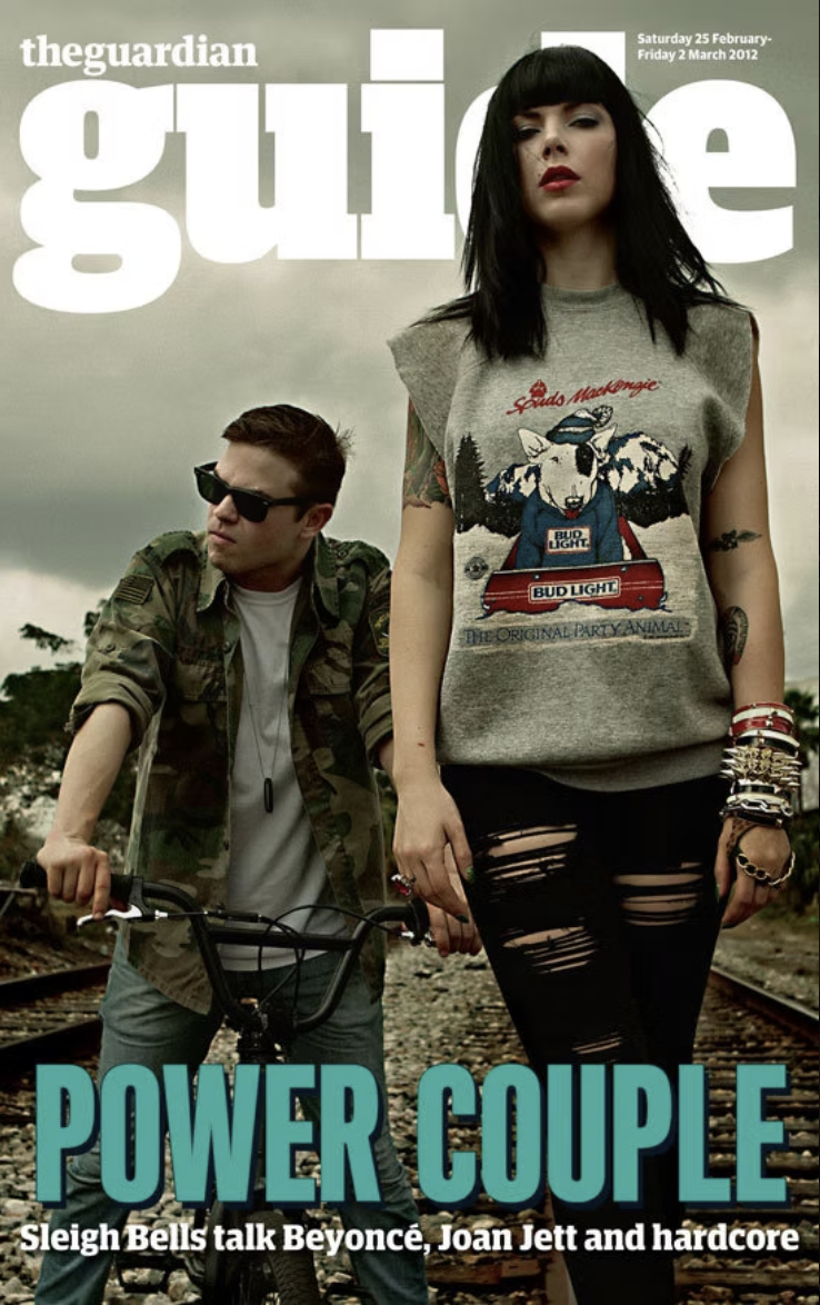 Cover of The Guardian Guide magazine featuring two young adults, a man and woman, standing on railway tracks. The woman has black hair, wears a gray sleeveless shirt with a graphic and several bracelets, and is looking at the camera. The man has shor