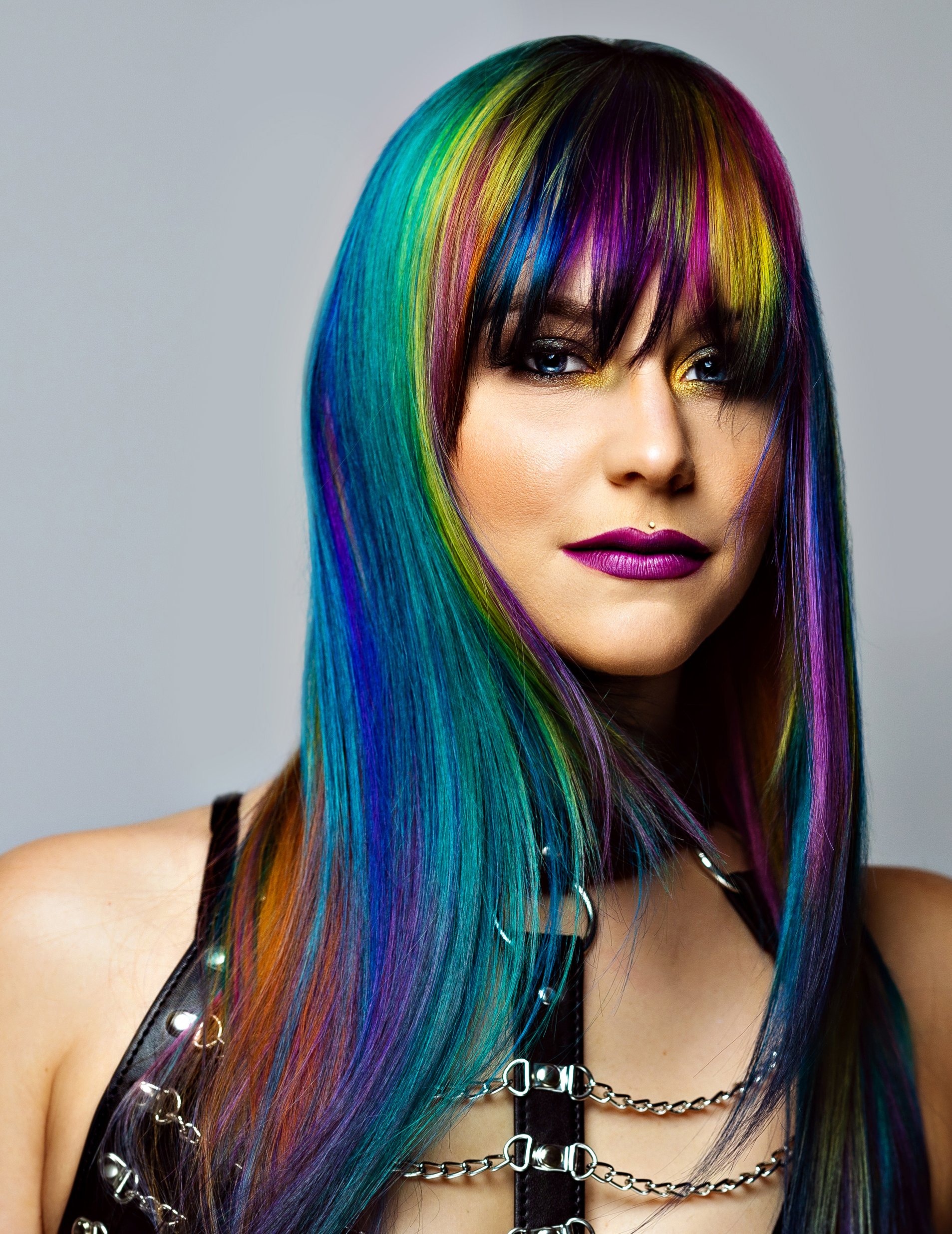 A woman with multi-colored hair including shades of rainbow, black, and dark purple, styled with layers and fringe, wearing makeup with gold eyeshadow,  a black leather top with metallic studs, and chains as accessories. hair tinsel near UCF