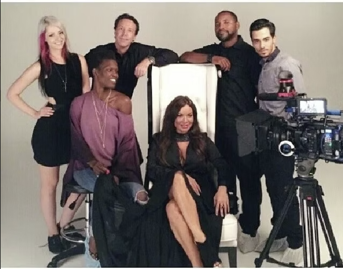 A diverse group of six people, four men and two women, poses for a photo behind a camera setup. One woman is sitting in a white chair at the center, and the others stand or sit around her, smiling. They are in a studio with a plain background.
