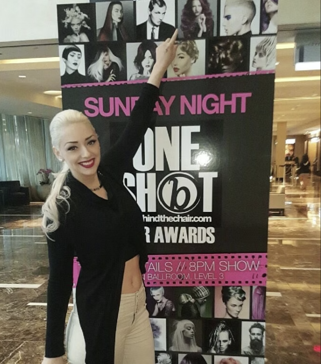 A young woman with platinum blonde hair styled in a high ponytail, wearing a black jacket and beige pants, smiling and pointing at a poster featuring various black and white and color images of women with different hairstyles, with a large text promo