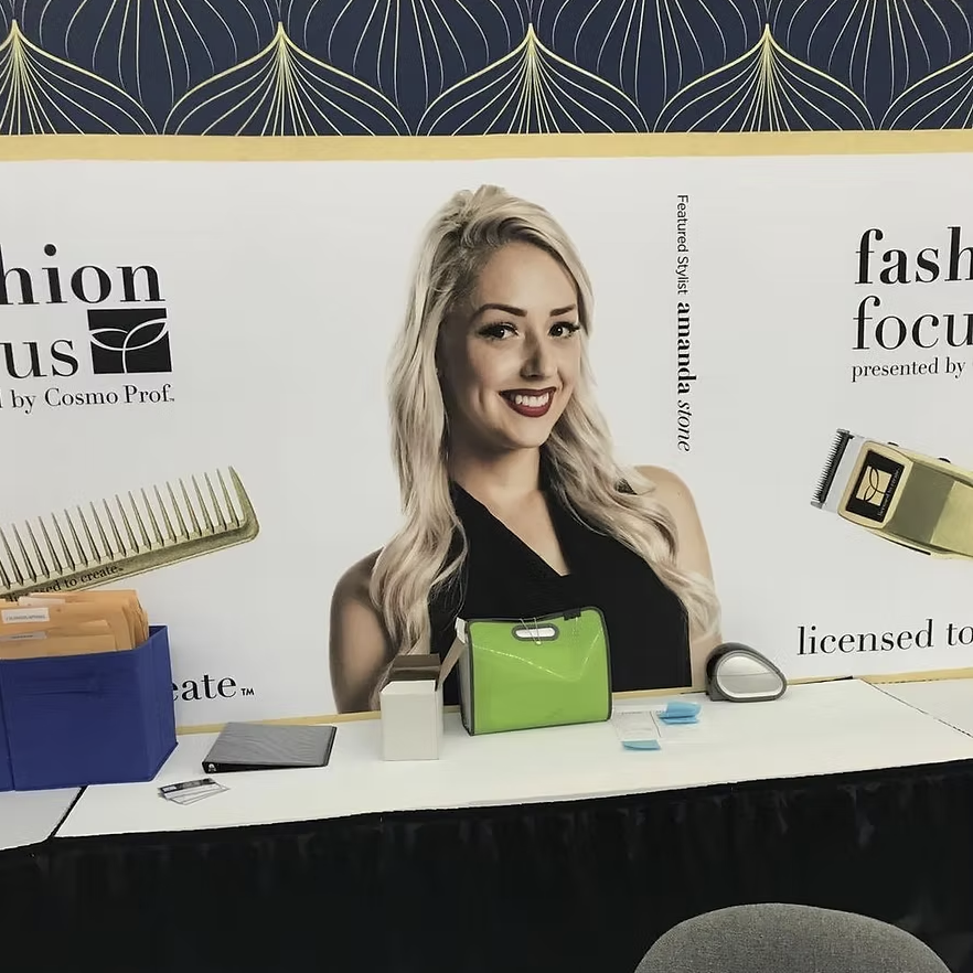 A woman with blonde hair wearing a black sleeveless top standing behind a white table with various office supplies in front of a backdrop that displays text and images related to hair styling, including scissors, a comb, and hair clippers.