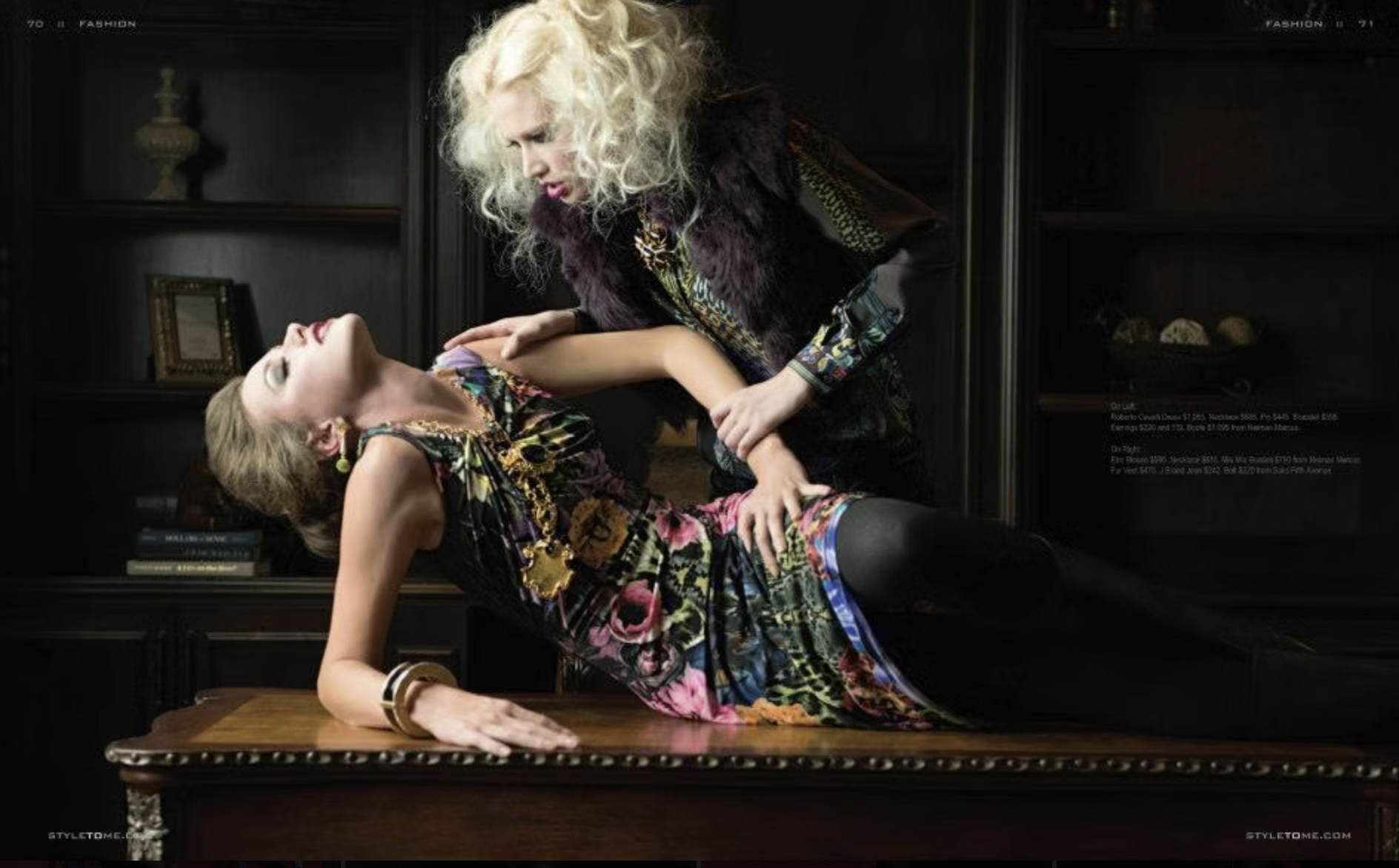 Two women in colorful fashion outfits in a dark room with bookshelf. One woman is sitting at a table, eyes closed, with a hand on the table. The other woman is leaning on her, with hands on her arm, looking at her.