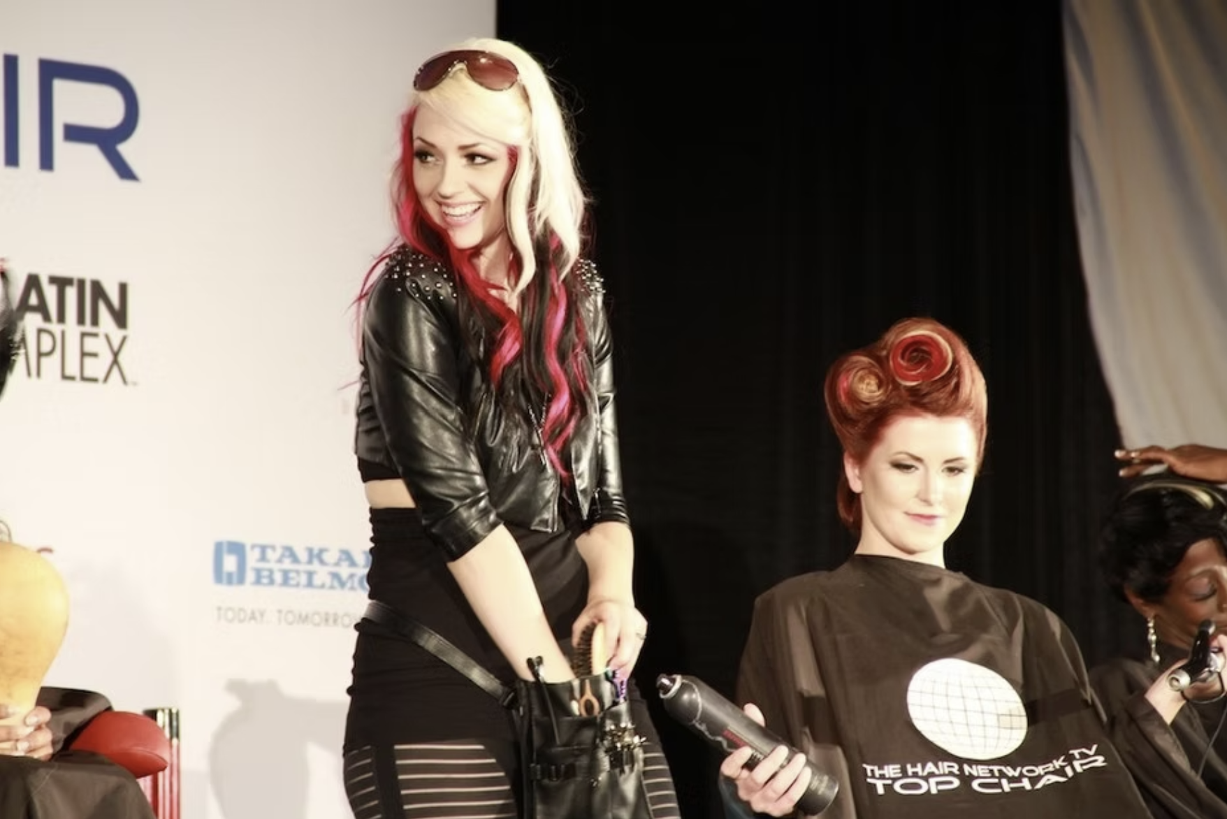 Woman with blonde and pink hair, wearing sunglasses on her head, styling the hair of another woman with vintage red curls, seated in a styling chair at a hair show or event.