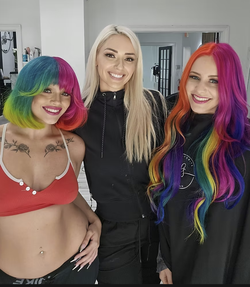 Three women smiling indoors, with the two on the sides having vibrant rainbow-colored hair, and the woman in the middle with long blonde hair, wearing a black jacket.