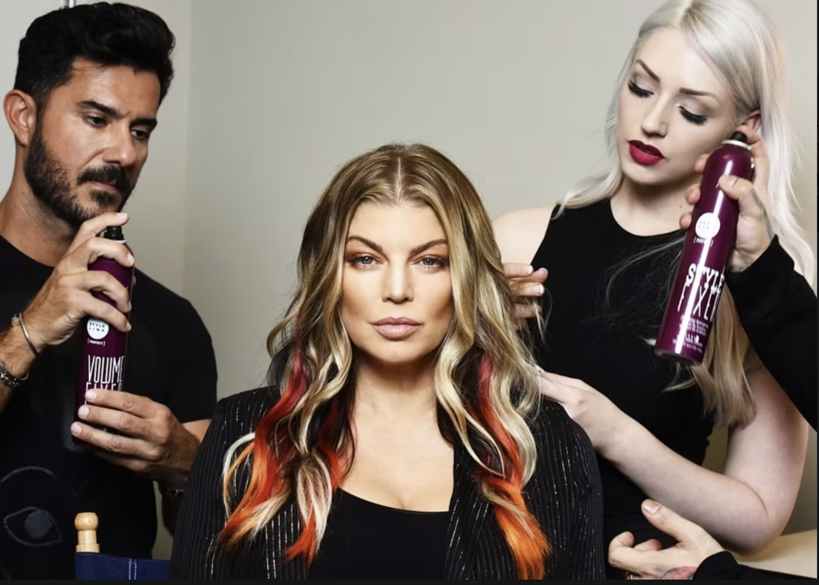 Hair stylists spray hair products onto a woman's long blonde hair with red highlights during a styling session.