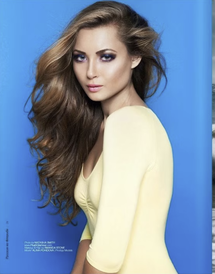 A woman with long, wavy brown hair and purple eyeshadow wearing a light yellow top against a blue background.