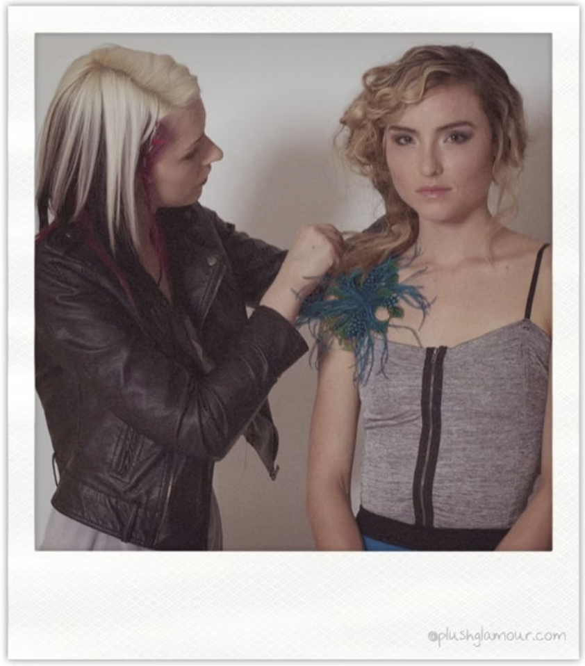 A hairstylist is working on a blonde woman's hair, which is styled in loose waves. The woman is wearing a grey top with black zipper details and a decorative blue flower adornment on her shoulder. The stylist has platinum blonde hair with pink streak