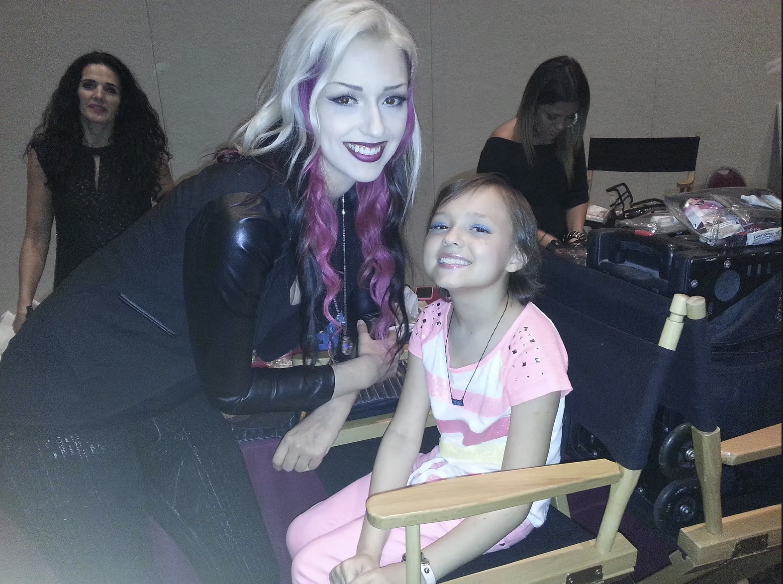 A woman with long, styled hair with purple streaks, wearing dark makeup and a black leather jacket, is smiling and leaning over a young girl sitting in a specialized chair. The girl has short brown hair, is smiling, wearing bright makeup, a pink and 