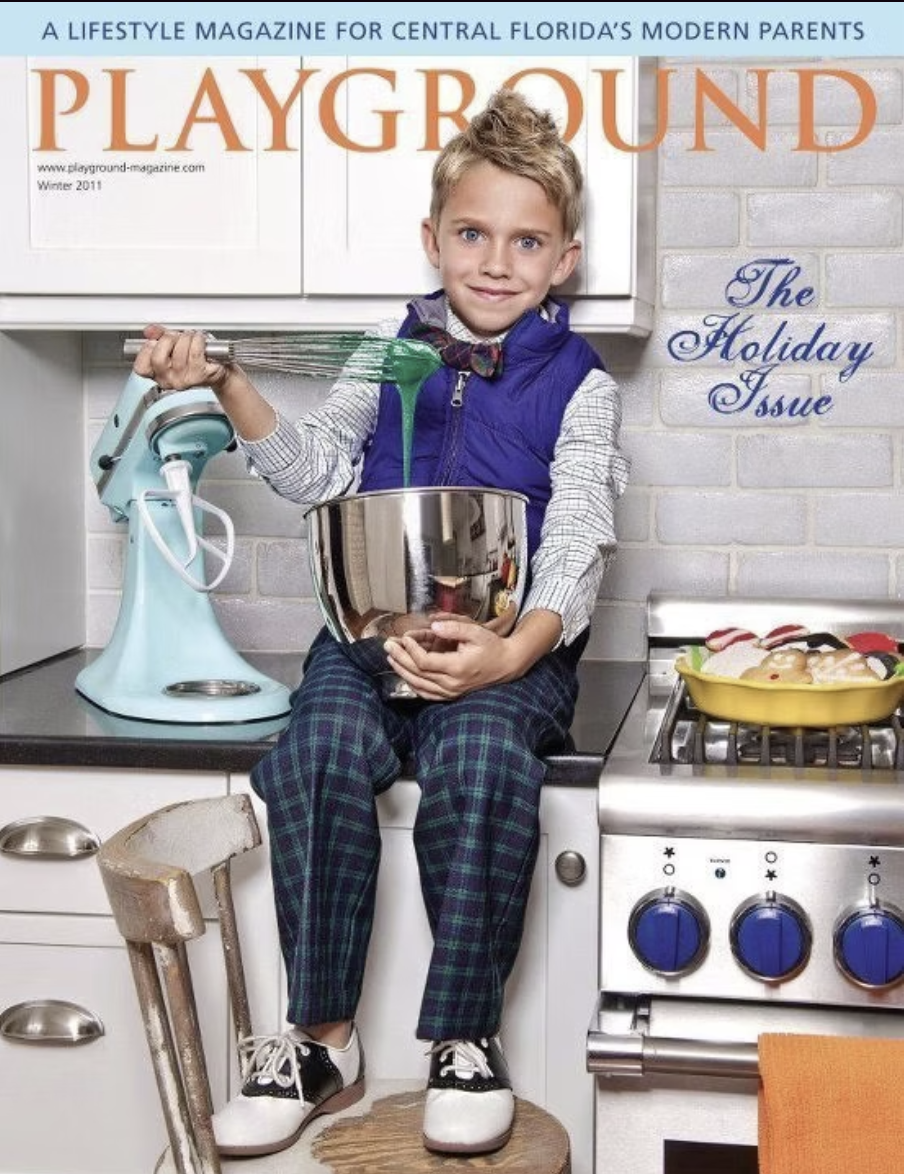 A young boy sitting on a kitchen counter holding a mixing bowl, with a hand mixer pouring green batter into the bowl. The kitchen has white cabinets, a stove with blue knobs, and a yellow dish of cookies. The magazine cover features the title 'Playgr