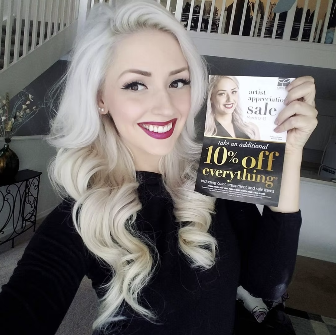 A woman with long, wavy platinum blonde hair wearing a black top smiling and holding a flyer promoting a sale with 10% off everything, including hair color, equipment, and sale items.