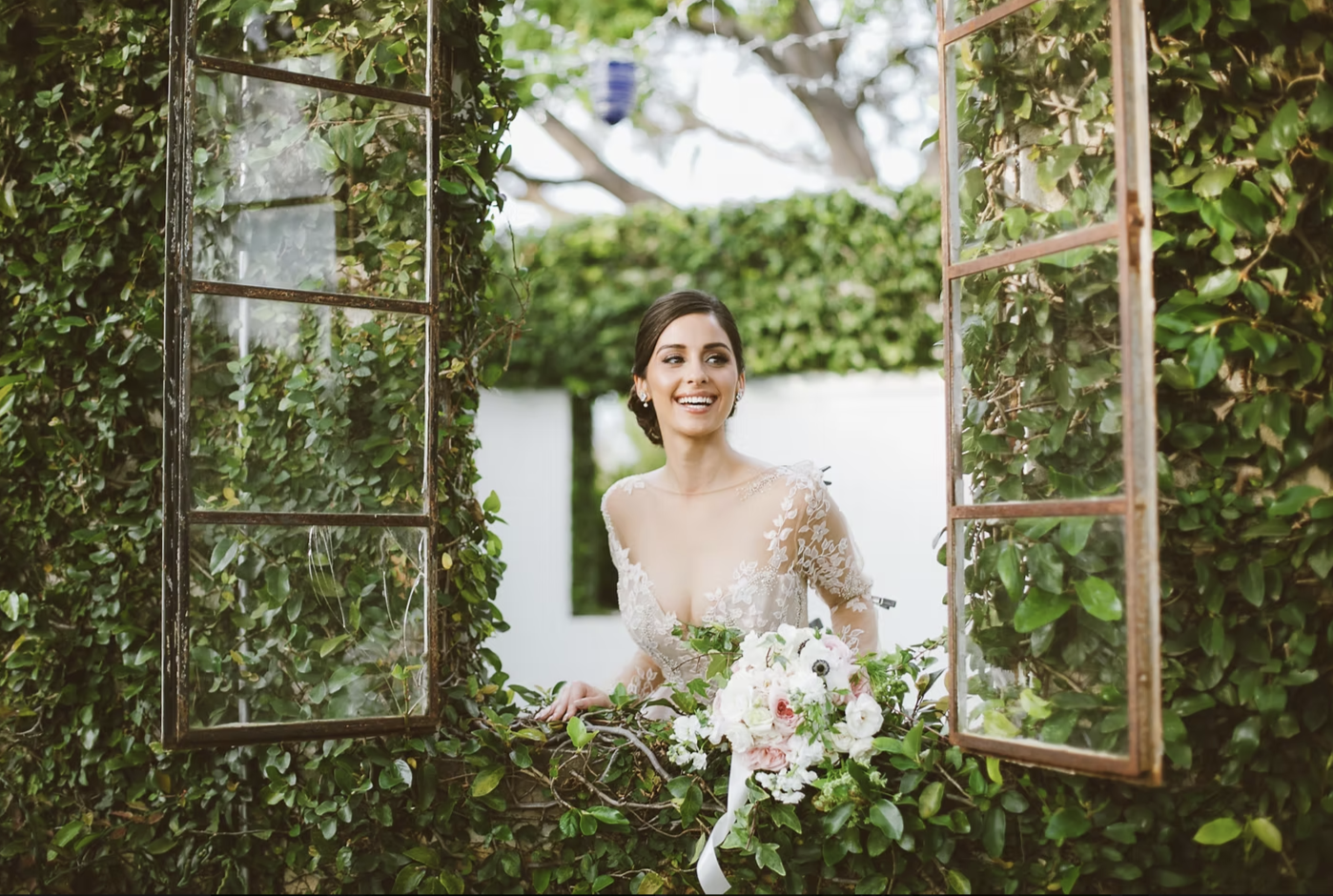 A woman in a wedding dress holding a bouquet of flowers, standing behind an open window surrounded by greenery. Bridal and wedding party services available at Salon Amoura near UCF and waterford lakes