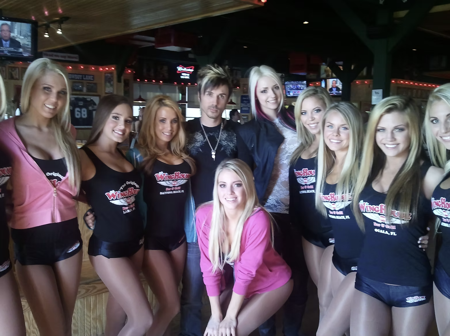 Group of women and a man posing for a photo inside a bar with wooden decor and TV screens, some women wearing black tank tops with 'Wingshoul' branding and one woman in a pink jacket, all smiling at the camera.