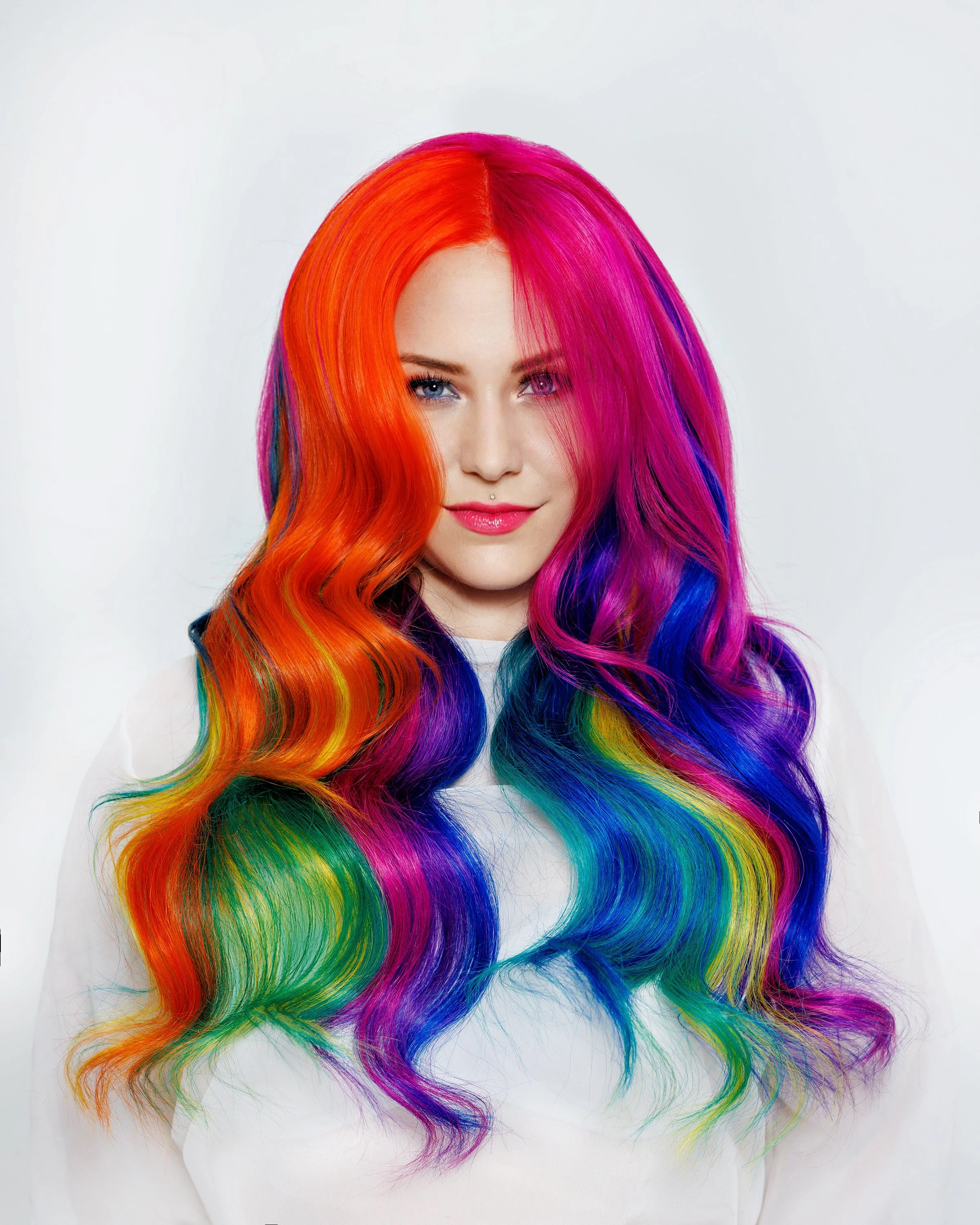 A woman with long, wavy hair dyed in a rainbow pattern including red, orange, yellow, green, blue, and purple, against a white background. Rainbow hair and tinsel located at Salon Amoura UCF avalon park