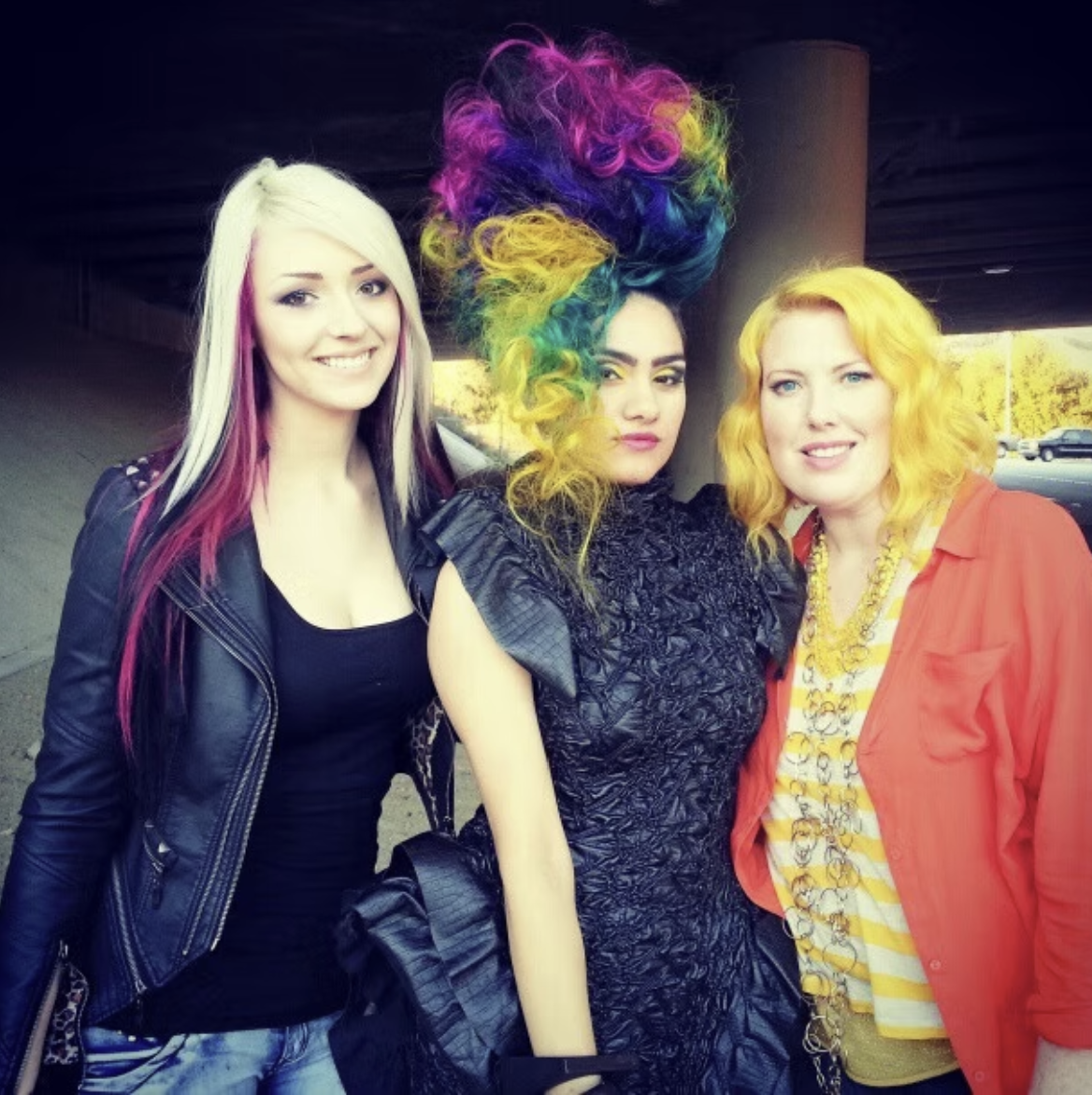 Three women standing together outdoors, one with rainbow-colored hair styled in an elaborate updo, and different fashionable clothing and hair colors.