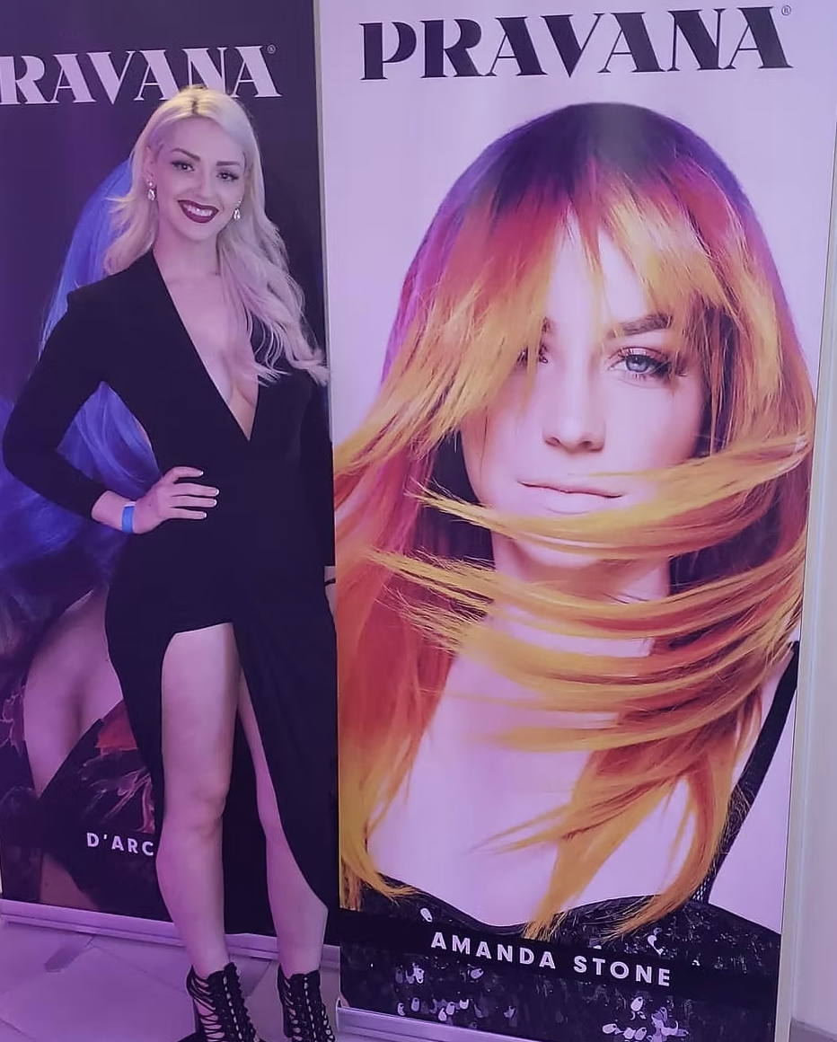 Woman standing next to large promotional posters for PRAVANA hair products featuring models with colorful hairstyles, one with blonde hair and the other with vibrant red and orange hair.