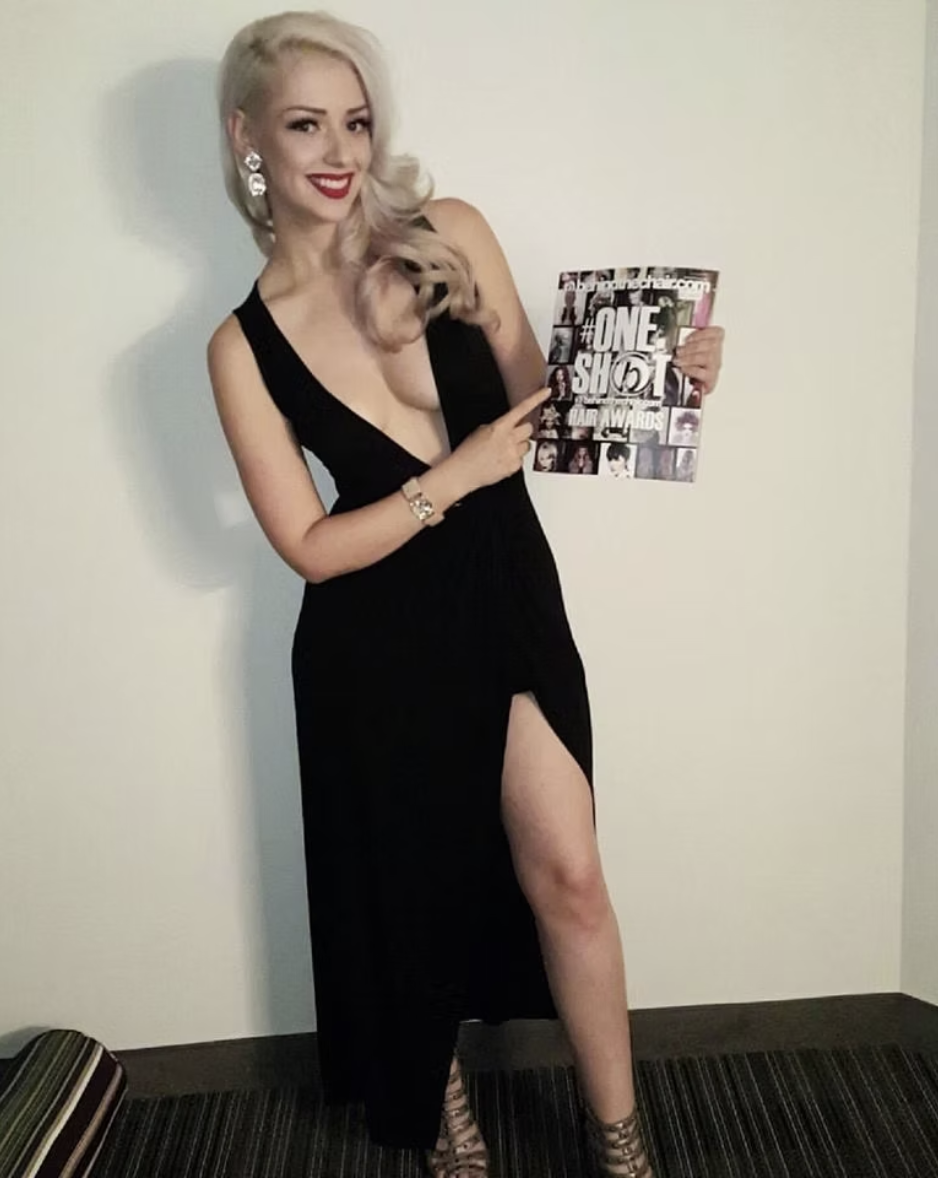 A woman with platinum blonde hair and red lipstick, wearing a black dress with a high slit, is smiling and pointing at a magazine she is holding. The magazine has bold text and images on its cover.