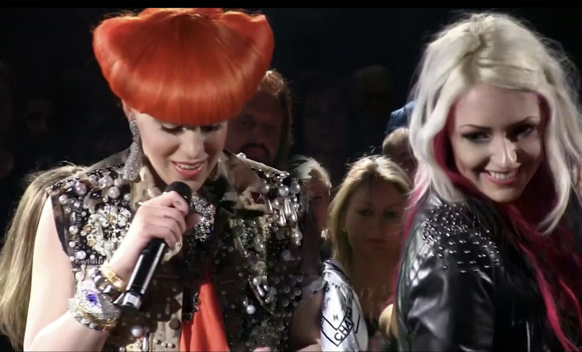 Two women with vibrant hair and dressed in studded jackets are on stage. One with orange hair is holding a microphone and smiling, the other has blonde hair with pink streaks and is also smiling.