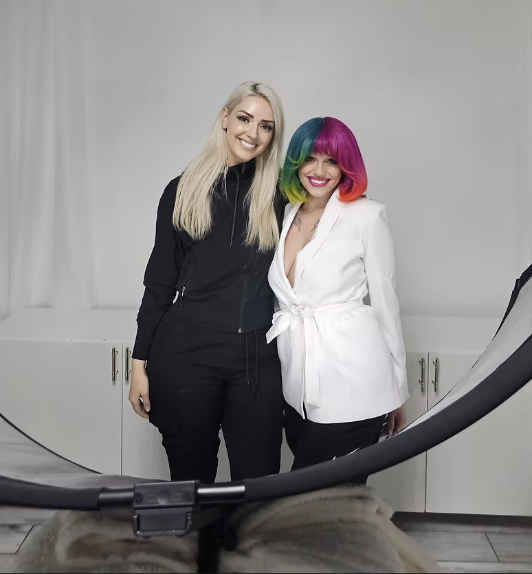 Two women standing close together, smiling at the camera. One has long blonde hair and is dressed in black, the other has a colorful, rainbow-hued bob hairstyle and is wearing a white belted blazer.