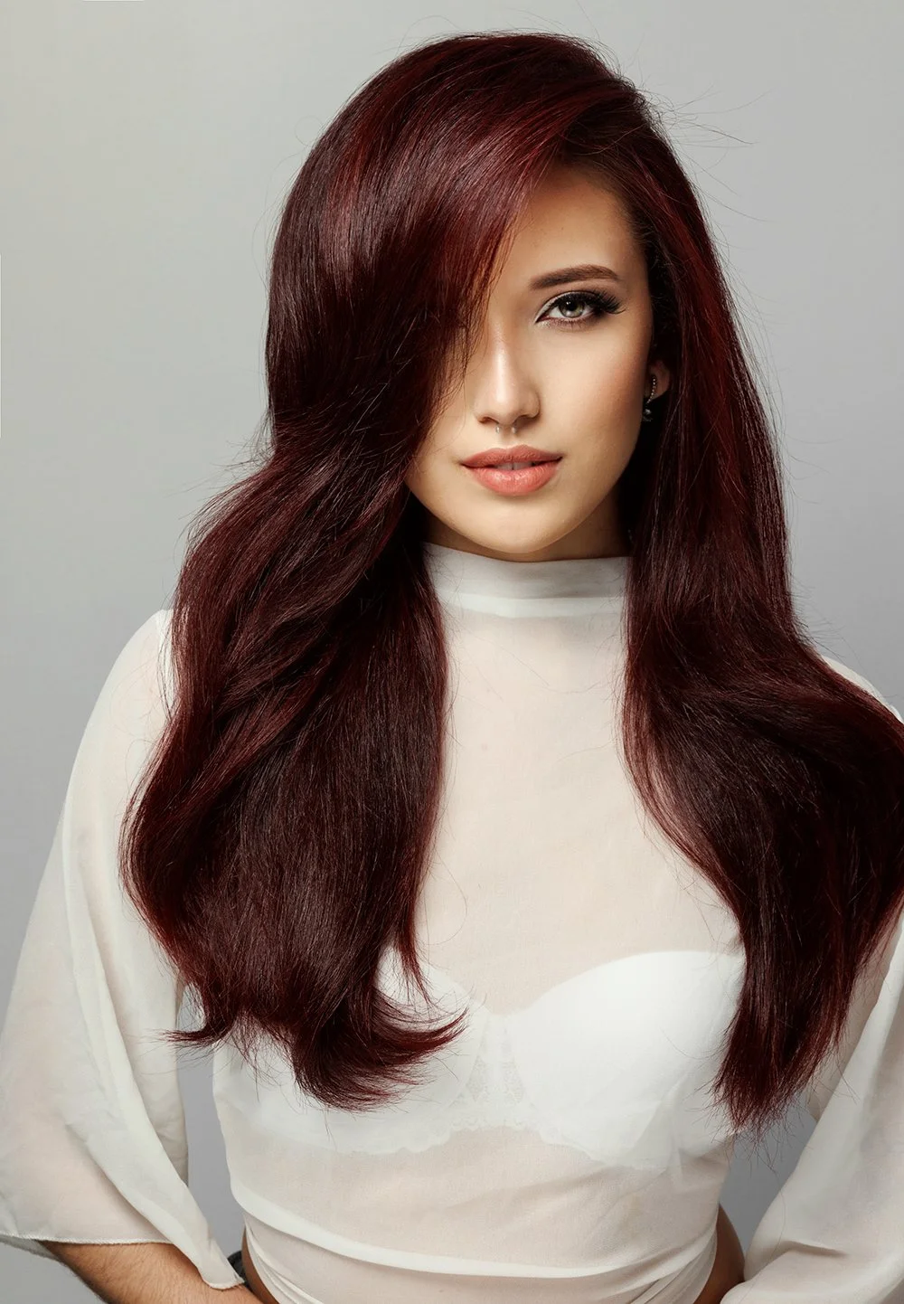 A woman with long, wavy, burgundy hair, wearing a white top, looking confidently at the camera against a plain light gray background. Brazilian Blowout done by Salon Amoura near UCF avalon park