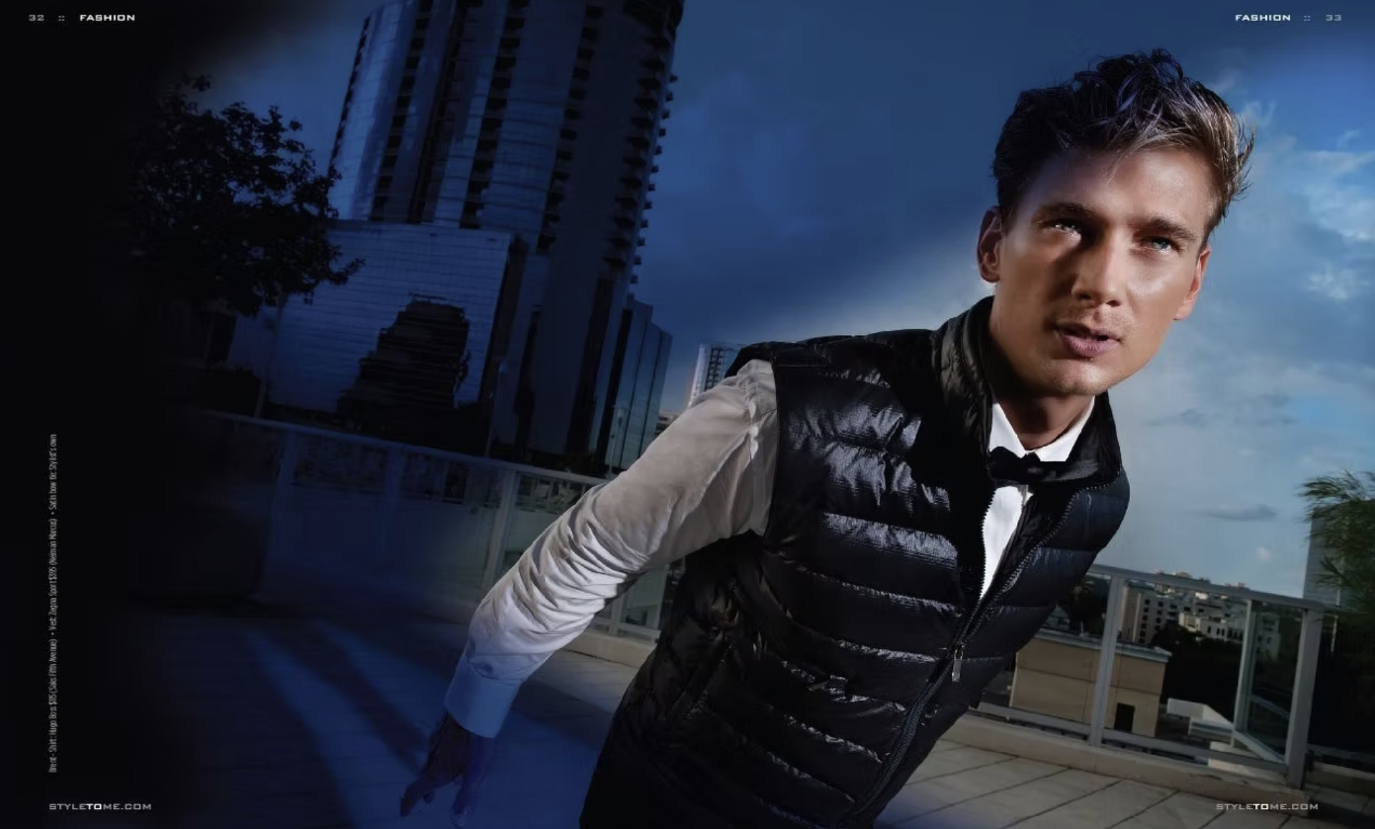 A young man in a black puffer vest over a white shirt standing outdoors on a rooftop with city buildings and a cloudy blue sky in the background.
