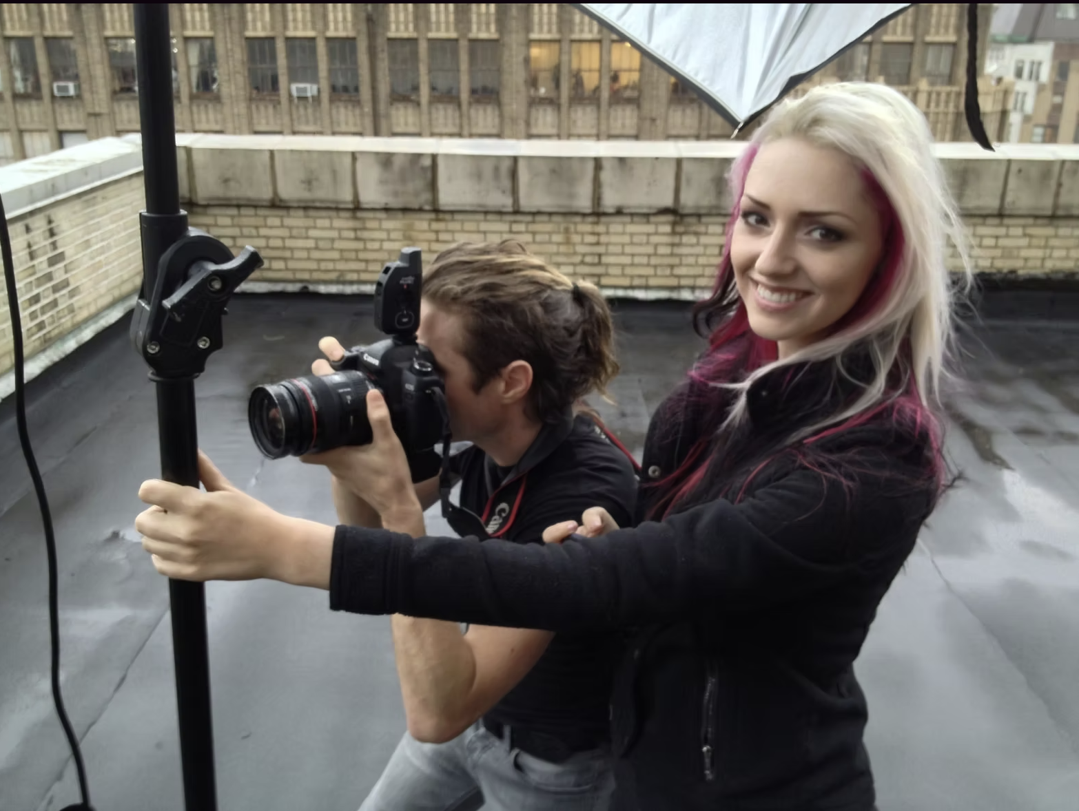 Two people on a rooftop, one woman smiling at the camera and a man taking a photo with a professional camera.