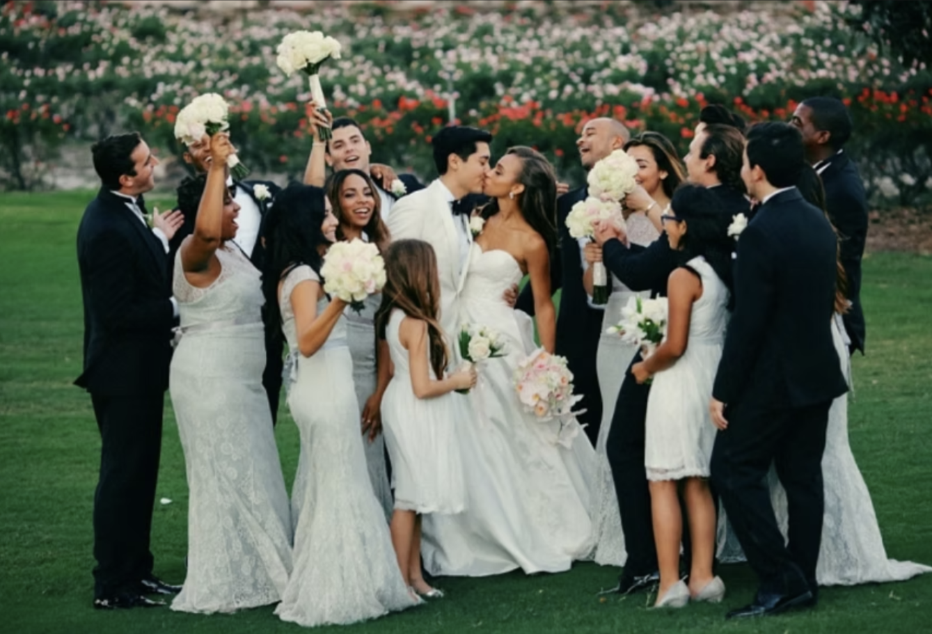 A wedding party outdoors on green grass with trees and flowers in background, showing the bride and groom kissing surrounded by bridesmaids and groomsmen holding bouquets and celebrating.
