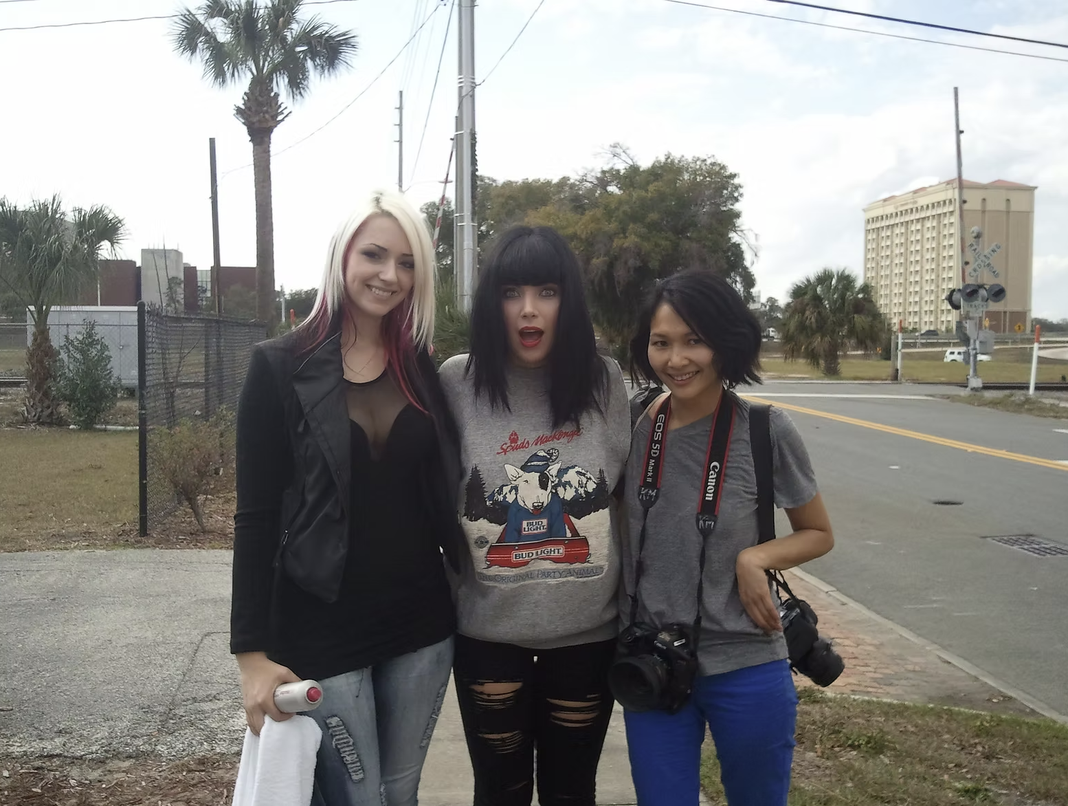 Three women standing outdoors on a sidewalk, smiling, with a Florida hotel in the background. One woman has blond hair with pink streaks, a black leather jacket, and gray pants. The second woman has black hair with bangs, wearing a gray sweatshirt wi