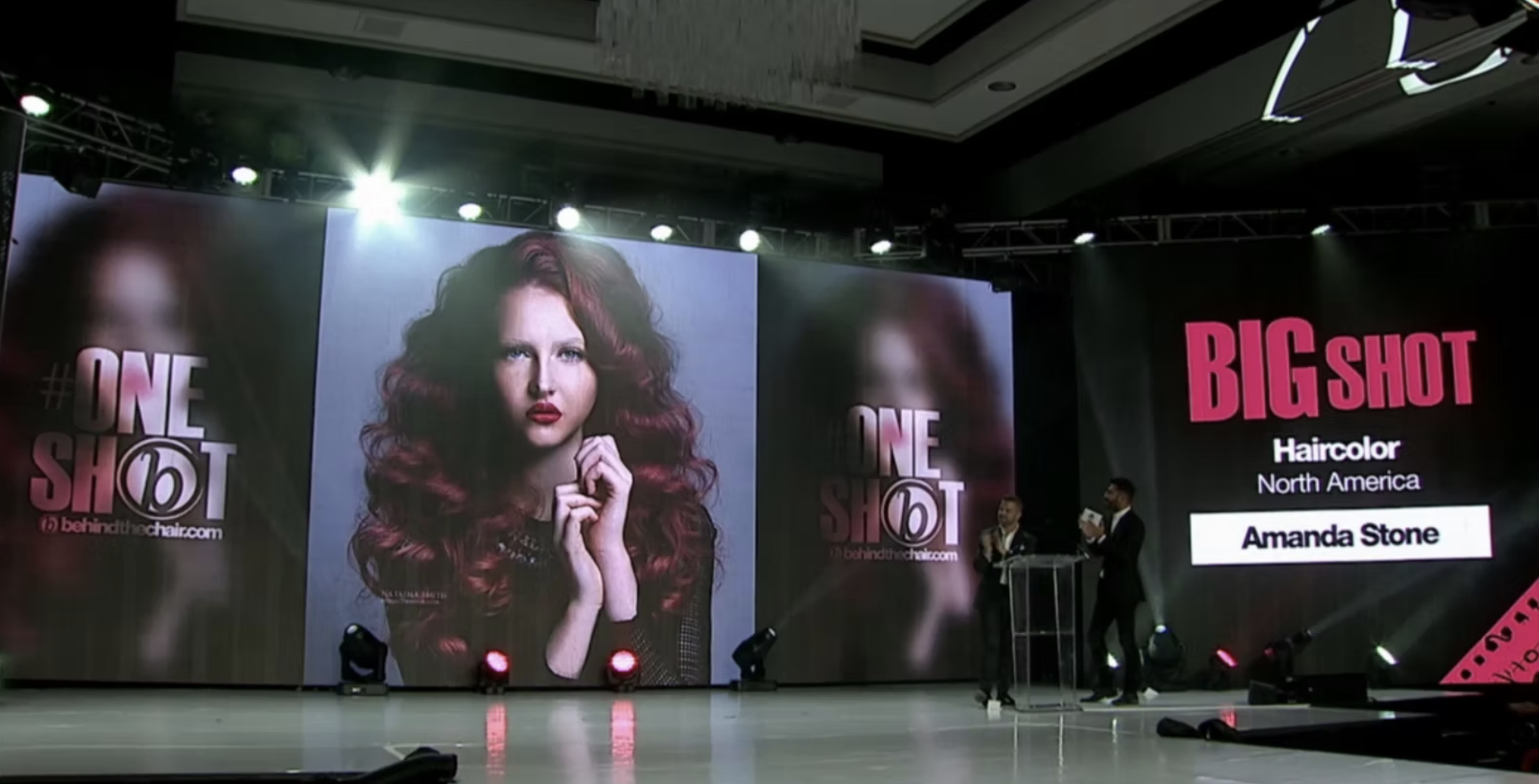 A stage with large screens displaying images of a woman with long, curly, red hair and the text 'ONE SHOT' and 'BIG SHOT'. The stage is lit with spotlights, and there are people in the background, including a person at a podium.