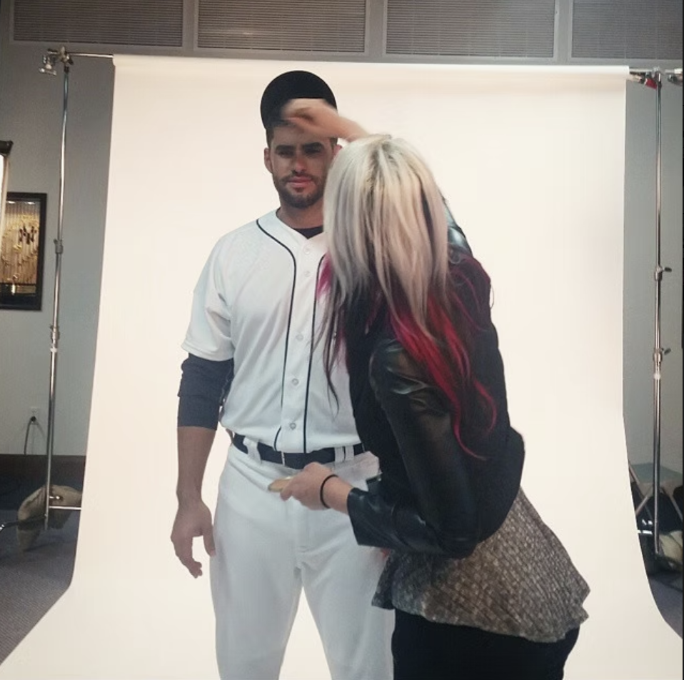 A person in a baseball uniform standing in front of a photo backdrop, with a woman taking notes or signing an autograph.