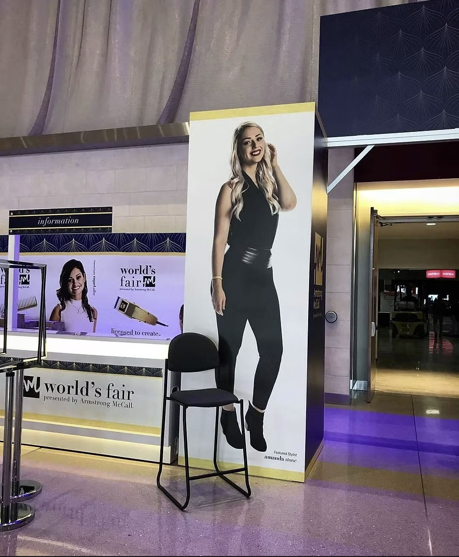 Large promotional display of a woman with long blonde hair wearing a black sleeveless top, black pants, and ankle boots, standing in front of a white and yellow background at a public venue.