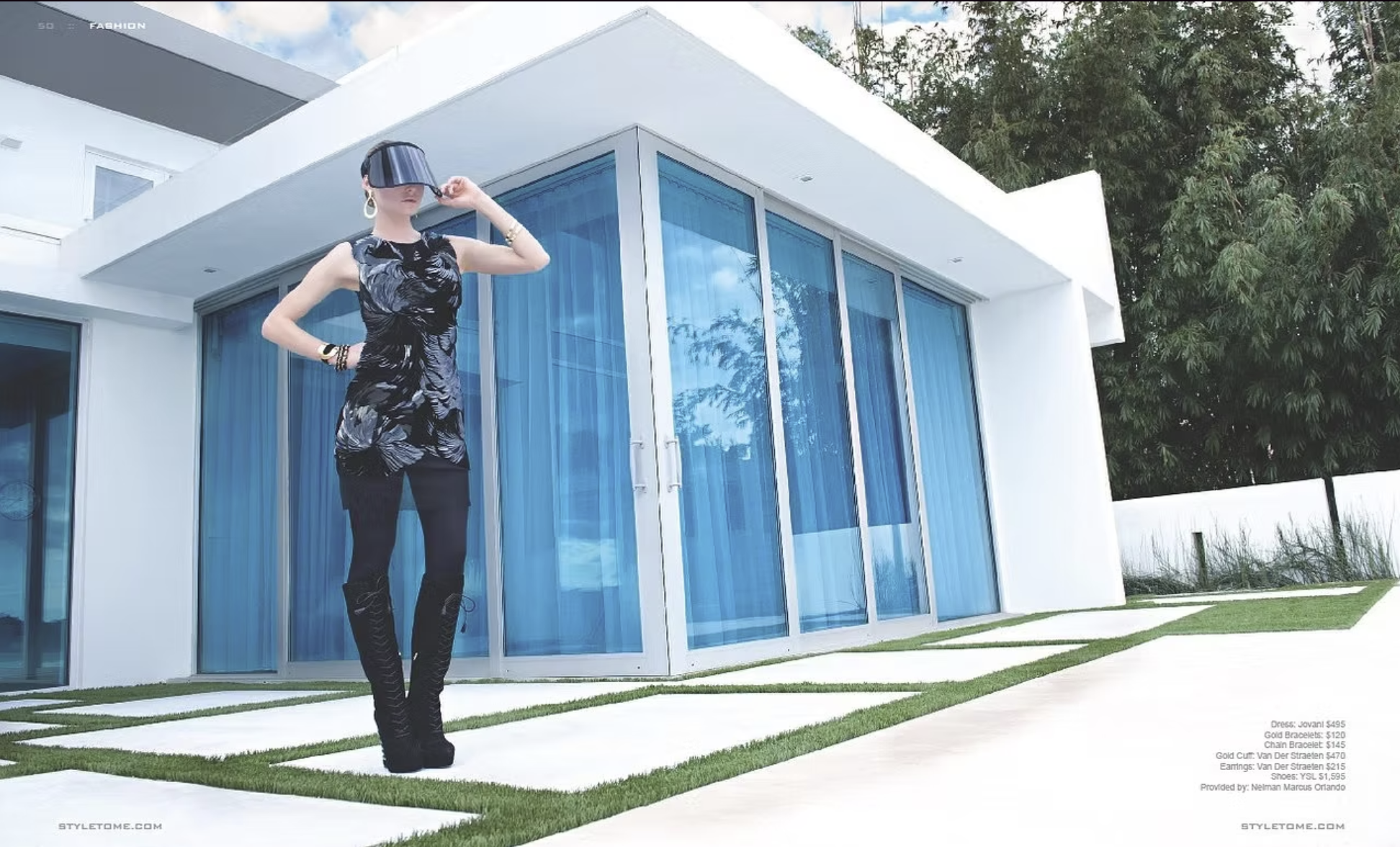 A woman standing outside a modern white house with large blue-tinted glass windows, wearing black pants, a black and gray patterned sleeveless dress, gold jewelry, and a black visor hat.