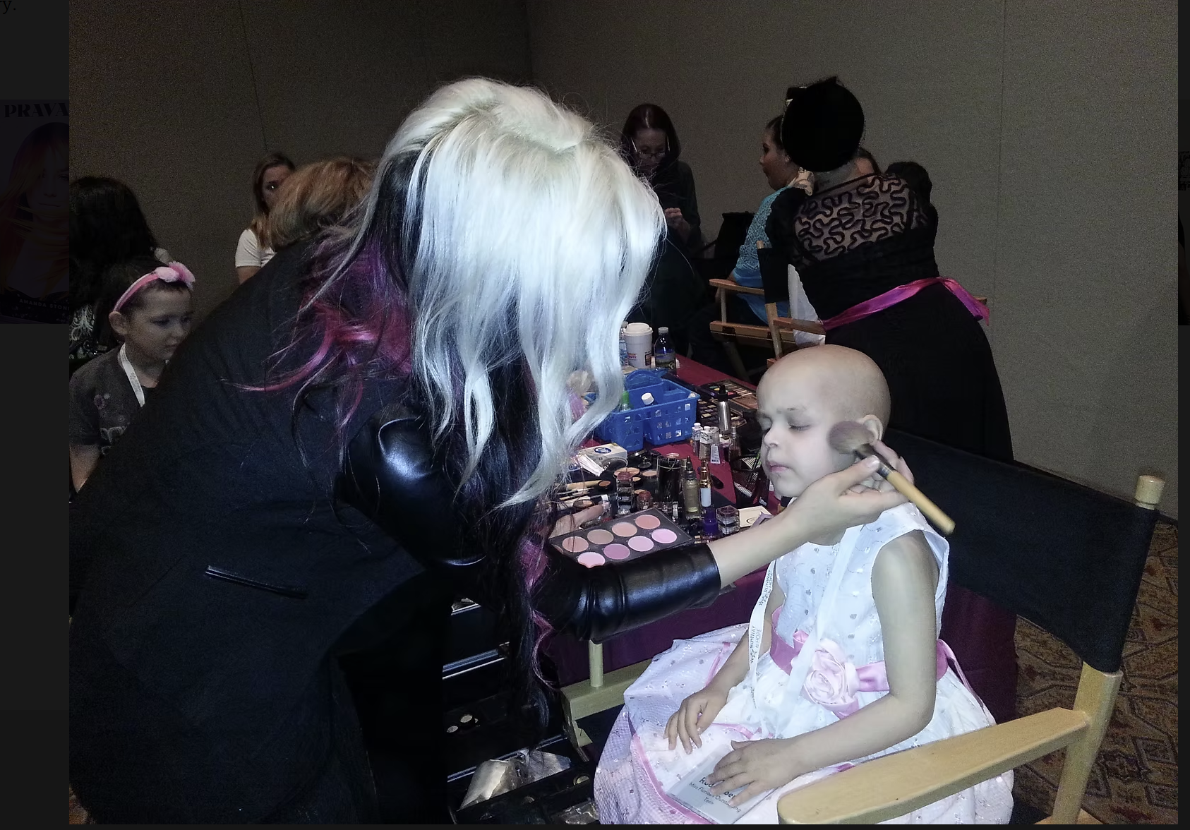 A young girl with a bald head, wearing a white dress with pink ribbons, sitting in a chair at a makeup station. A woman with colorful hair is applying makeup with a brush to the girl. In the background, several people are visible, including another w