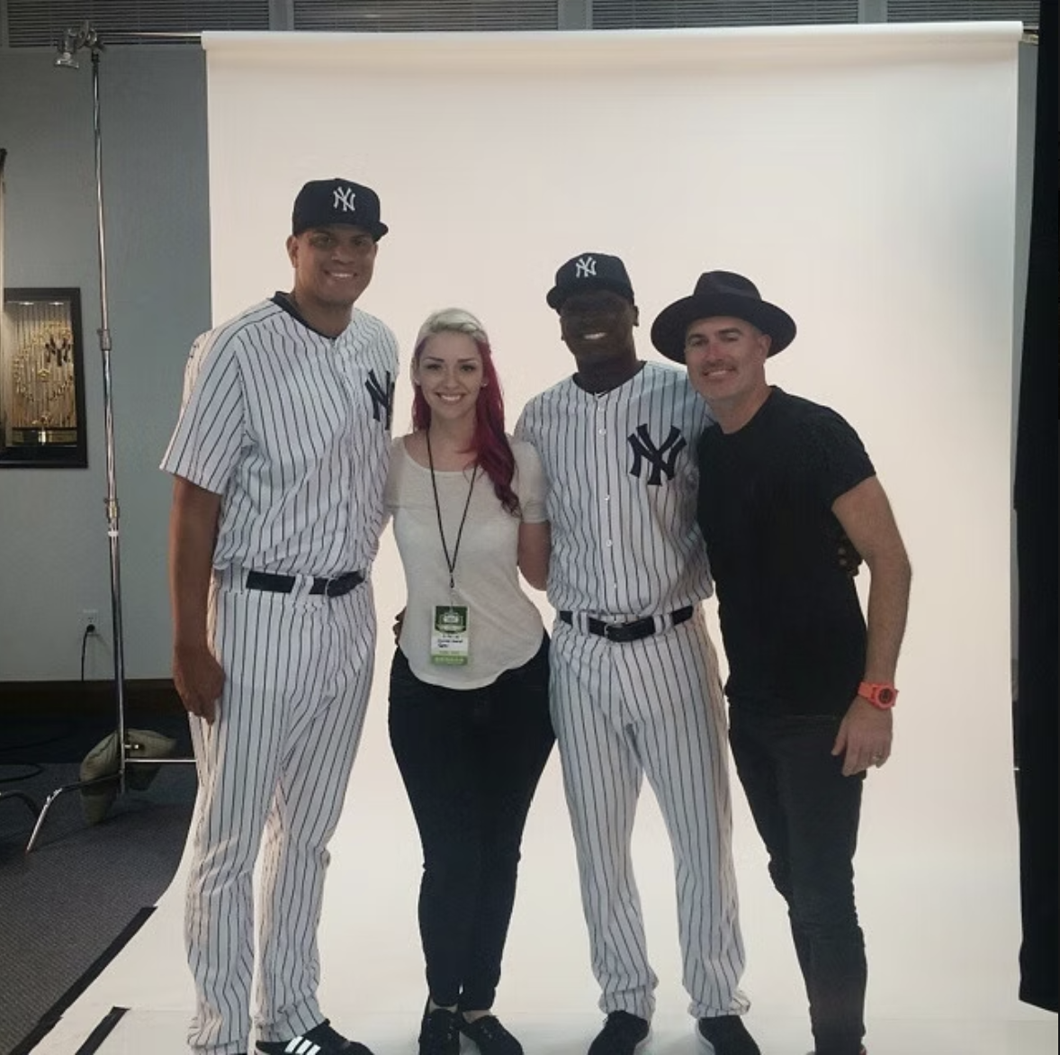 A group of four people standing in front of a white backdrop, with two individuals dressed as New York Yankees baseball players and two casually dressed people, one woman with pink hair and one man wearing a black hat and black shirt.