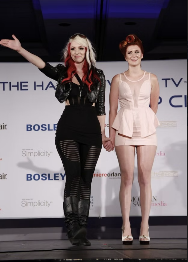 Two women on stage holding hands, with a backdrop displaying various logos and text including 'The Hair', 'Bosley', and 'Simplicity'. One woman has blonde and red hair, wearing a black outfit with a black leather jacket, and the other woman has red h