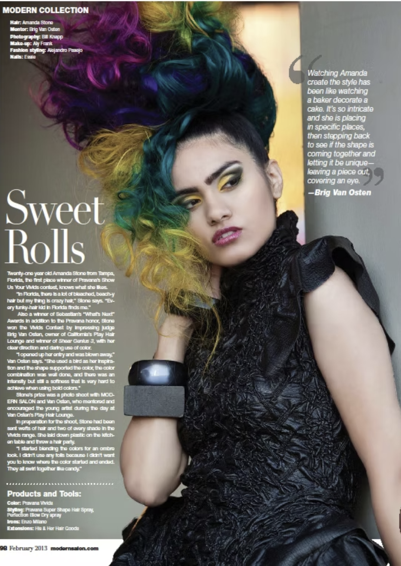 Young woman with colorful, curly hair in shades of purple, green, yellow, and blue, styled in an elaborate updo. She is wearing dark, dramatic makeup with yellow eyeshadow and dark lipstick, and a black textured sleeveless top. She has a large chunky