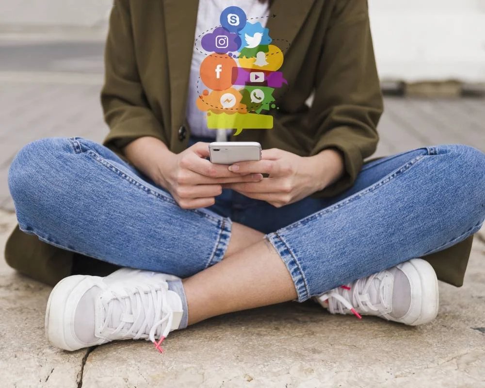 Person sitting cross-legged on a concrete surface, using a smartphone with social media icons emerging from the screen.